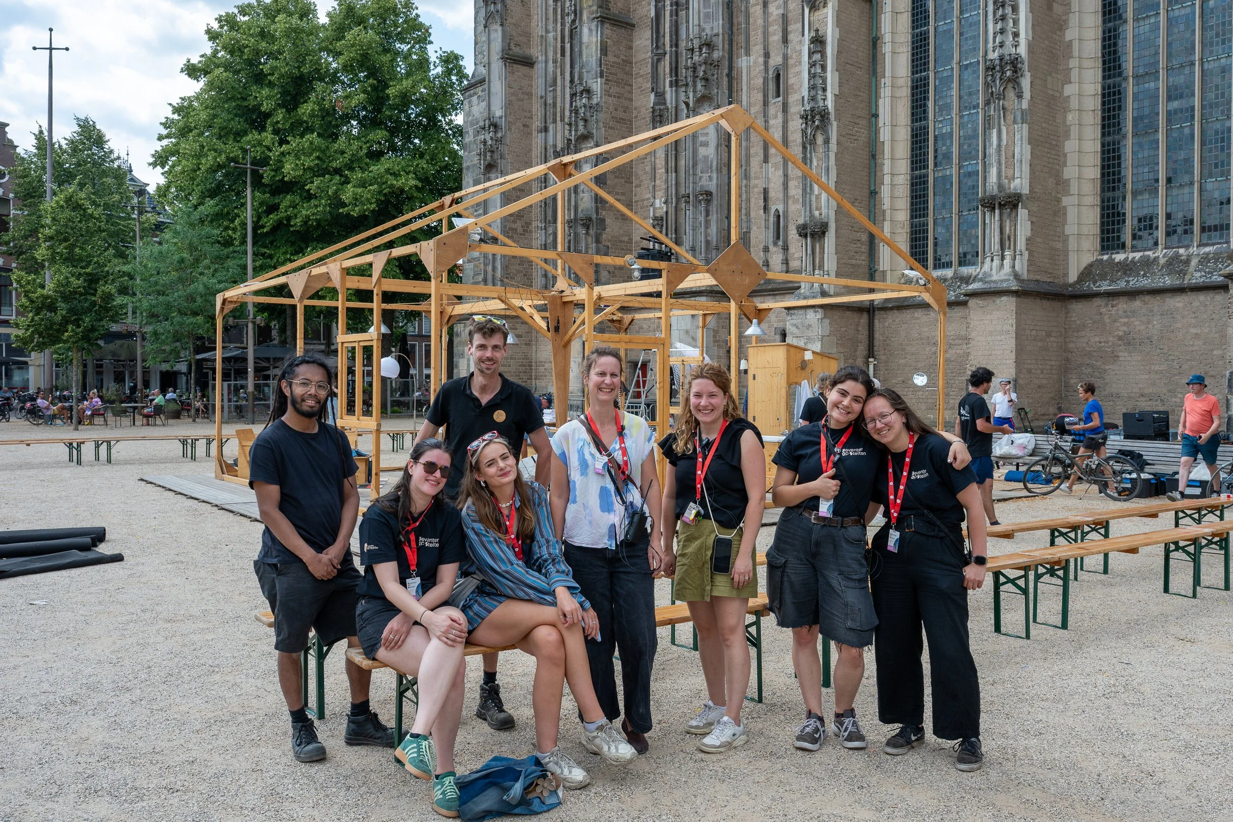 een groep jonge mensen die voor een houten frame buitenshuis staan, met een historische kerk op de achtergrond.