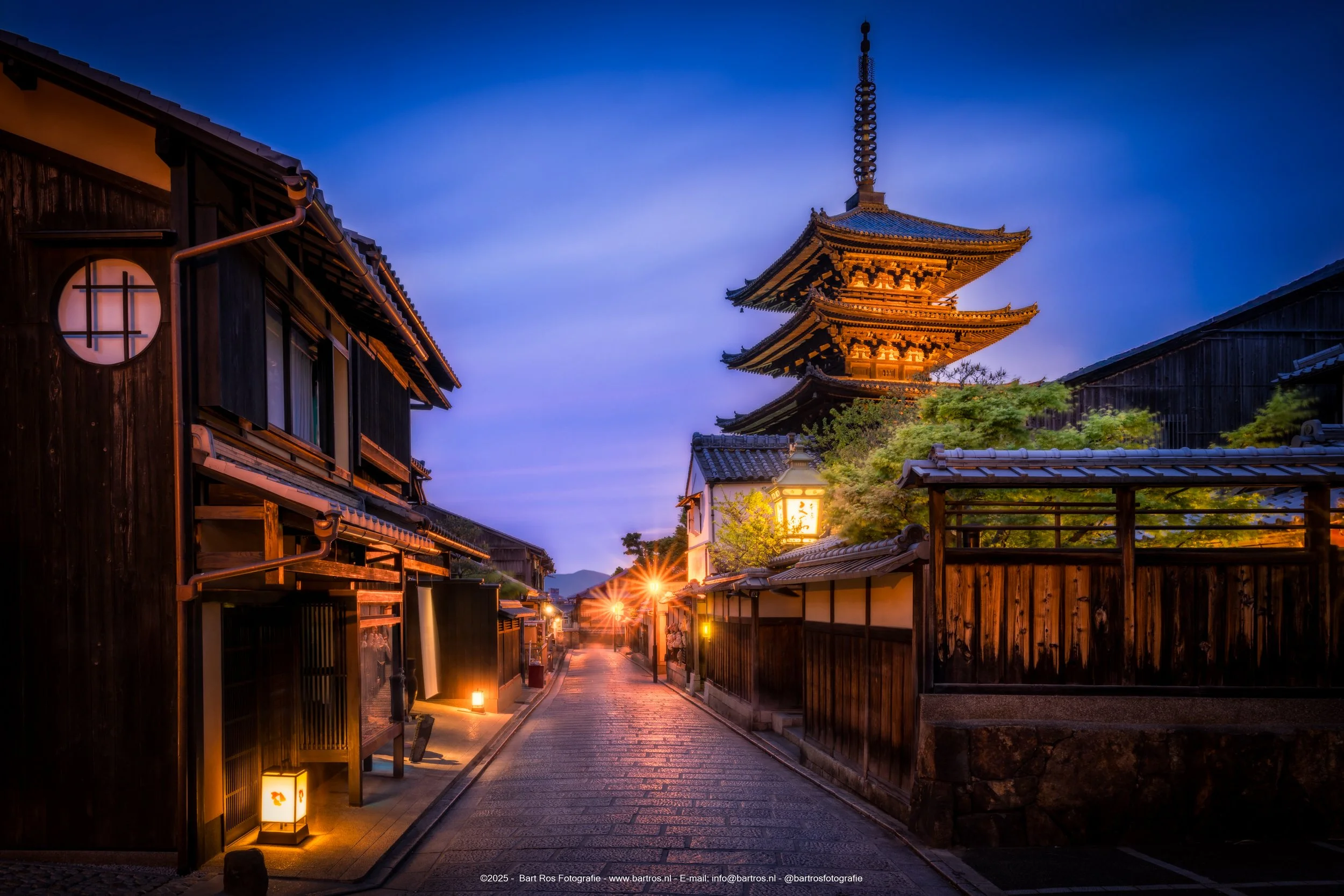 Een straat in Japan met traditionele houten gebouwen en een pagodeturm in de achtergrond, verlicht door straatlampen 's avonds.