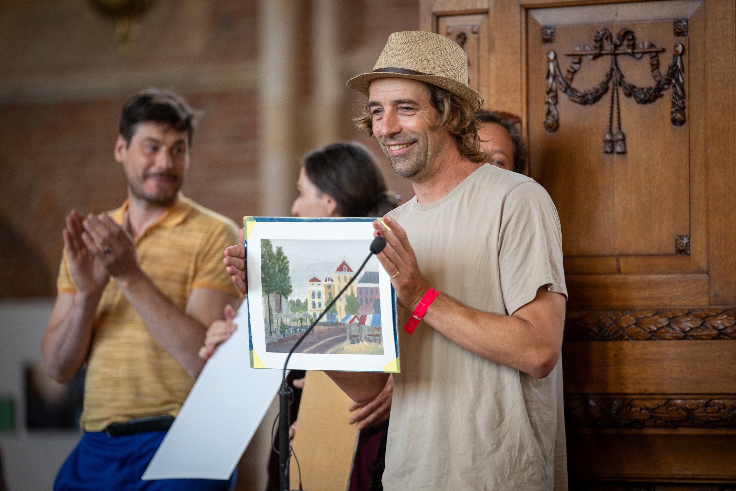 Man met een hoed die een tekening vasthoudt tijdens een presentatie, met mensen die applaudisseren in de achtergrond.