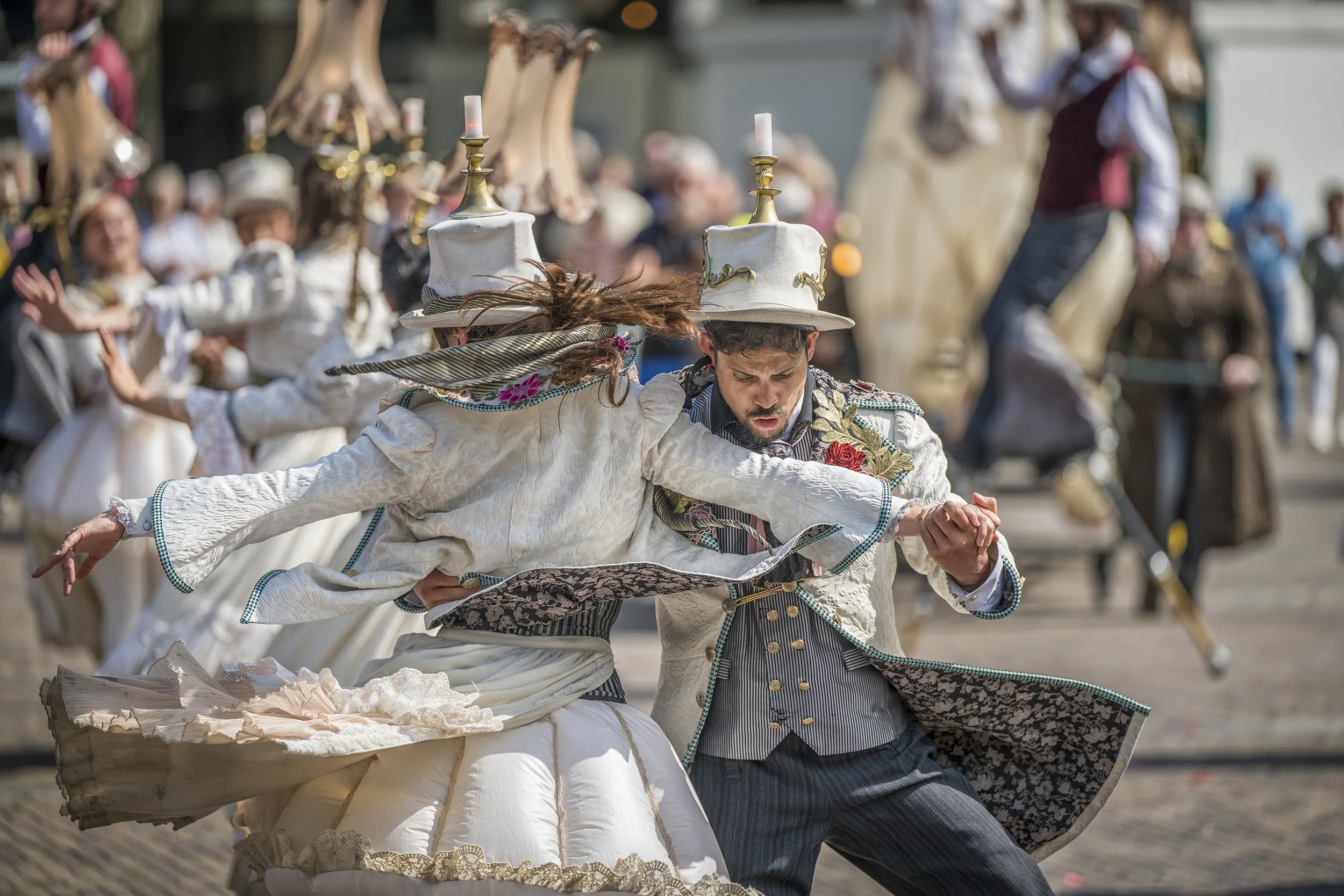 Mensen die deelnemen aan een traditionele volksdans, gekleed in elegante historische kostuums met hoeden en details, dansen in een parade of festival, met een publiek op de achtergrond.