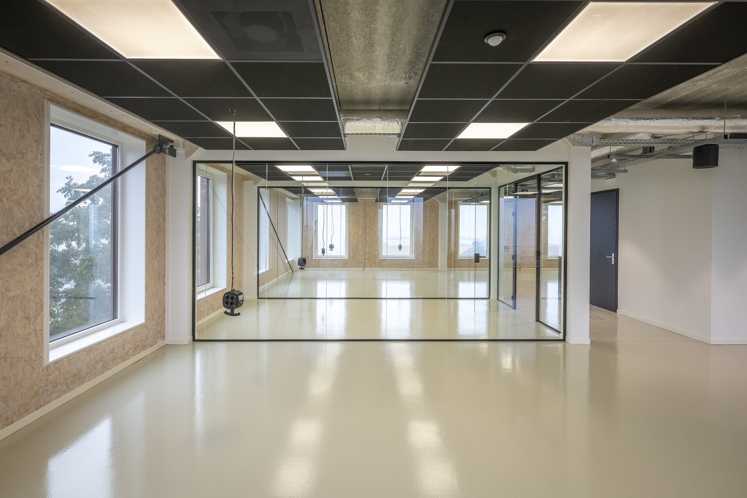 Een lege kamer met grote ramen, een spiegelwand, een plafond met zwarte en witte panelen en blootgelegde leidingwerken.