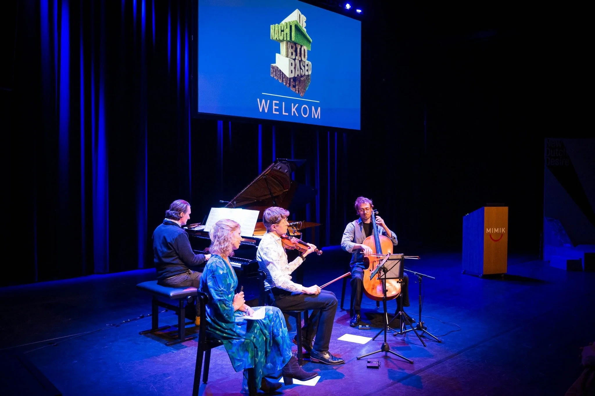 Muziekoptreden met vijf mensen die instrumenten bespelen op een podium in een donkere ruimte met blauwe verlichting en een groot scherm erboven dat 'Nacht van de Bio Based' en 'Welkom' toont.