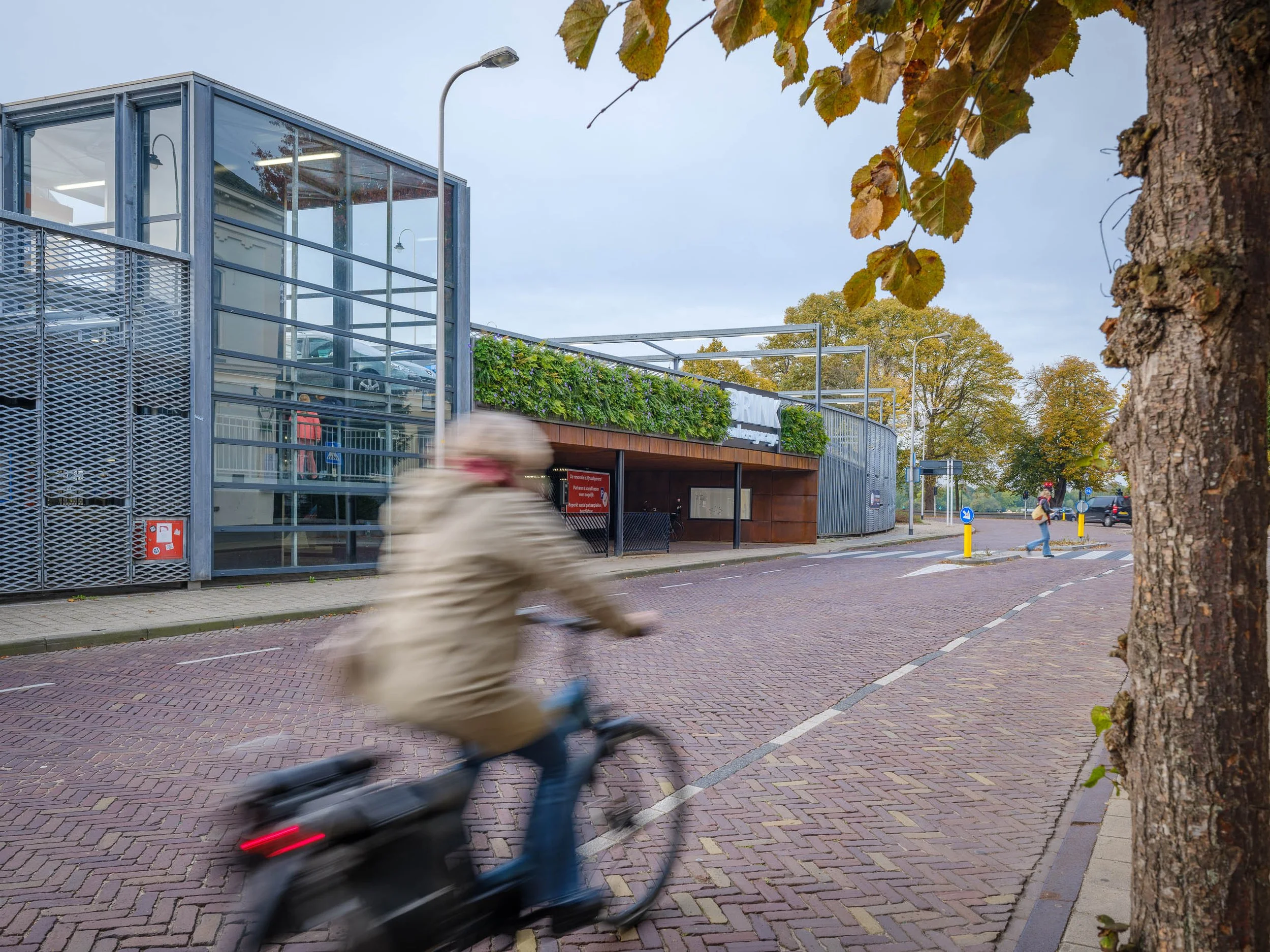 Een moderne glazen en metalen parkeergebouw met omringende bomen en een pavésstraat, terwijl een kind op een kleine fiets voorbij rijdt.