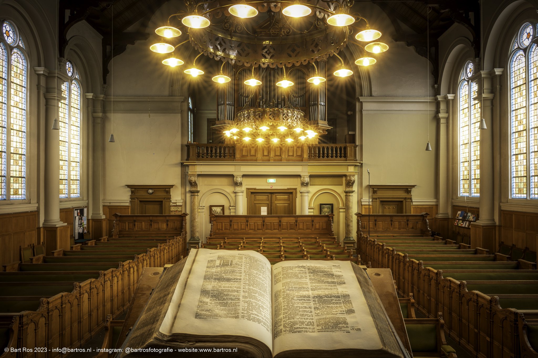 Binnenkant van een kerk met grote ramen, houten banken en een orgel op de achtergrond. In het midden een groot open boek op een tafel.