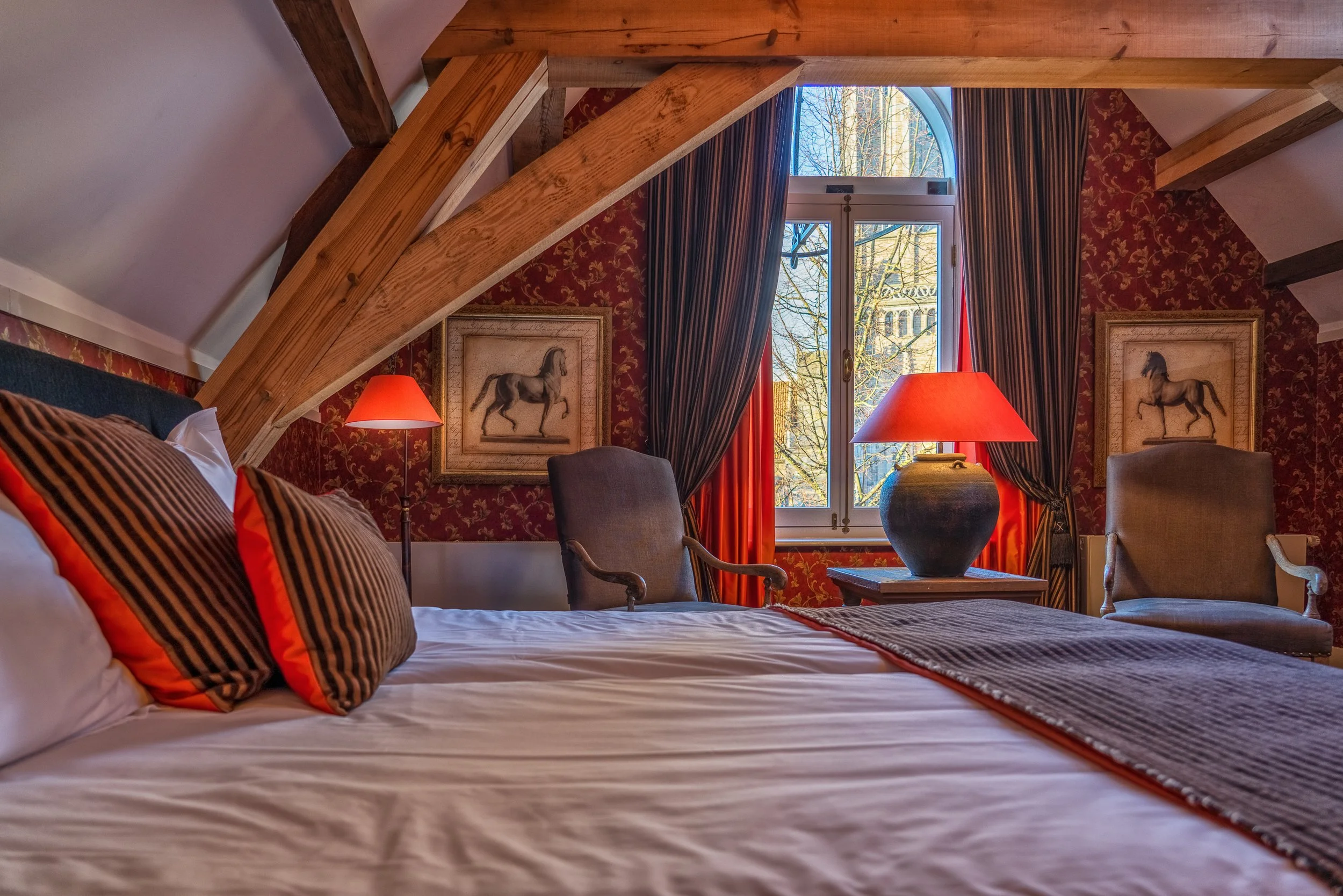 Slaapkamer met rood behang, een groot raam met uitzicht op bomen, twee lampen met oranje kappen, en ingelijste paardenafdrukken aan de muur.