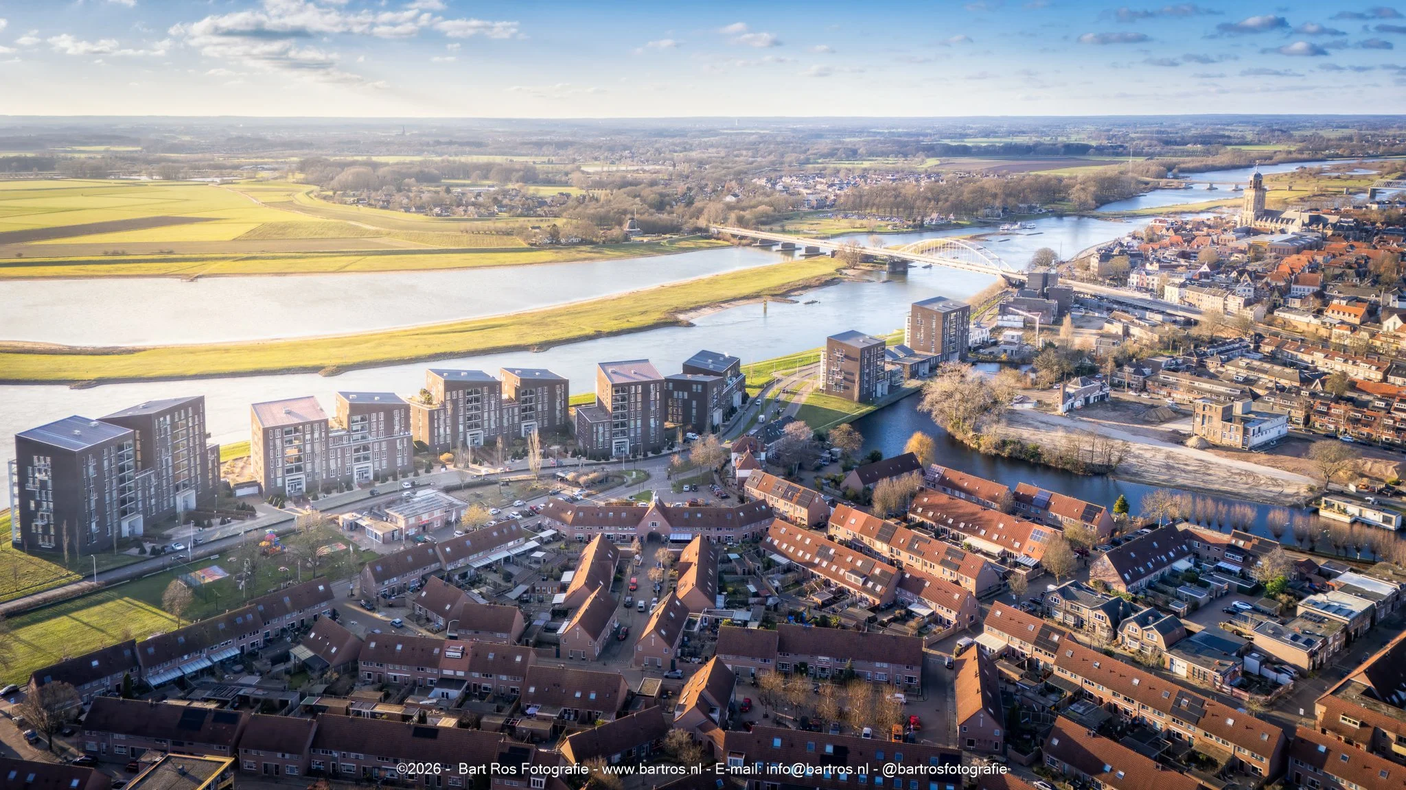 Luchtfoto van een stadsgebied met woonwijken, een rivier met bruggen, en groene velden op de achtergrond onder een partly bewolkte blauwe hemel.