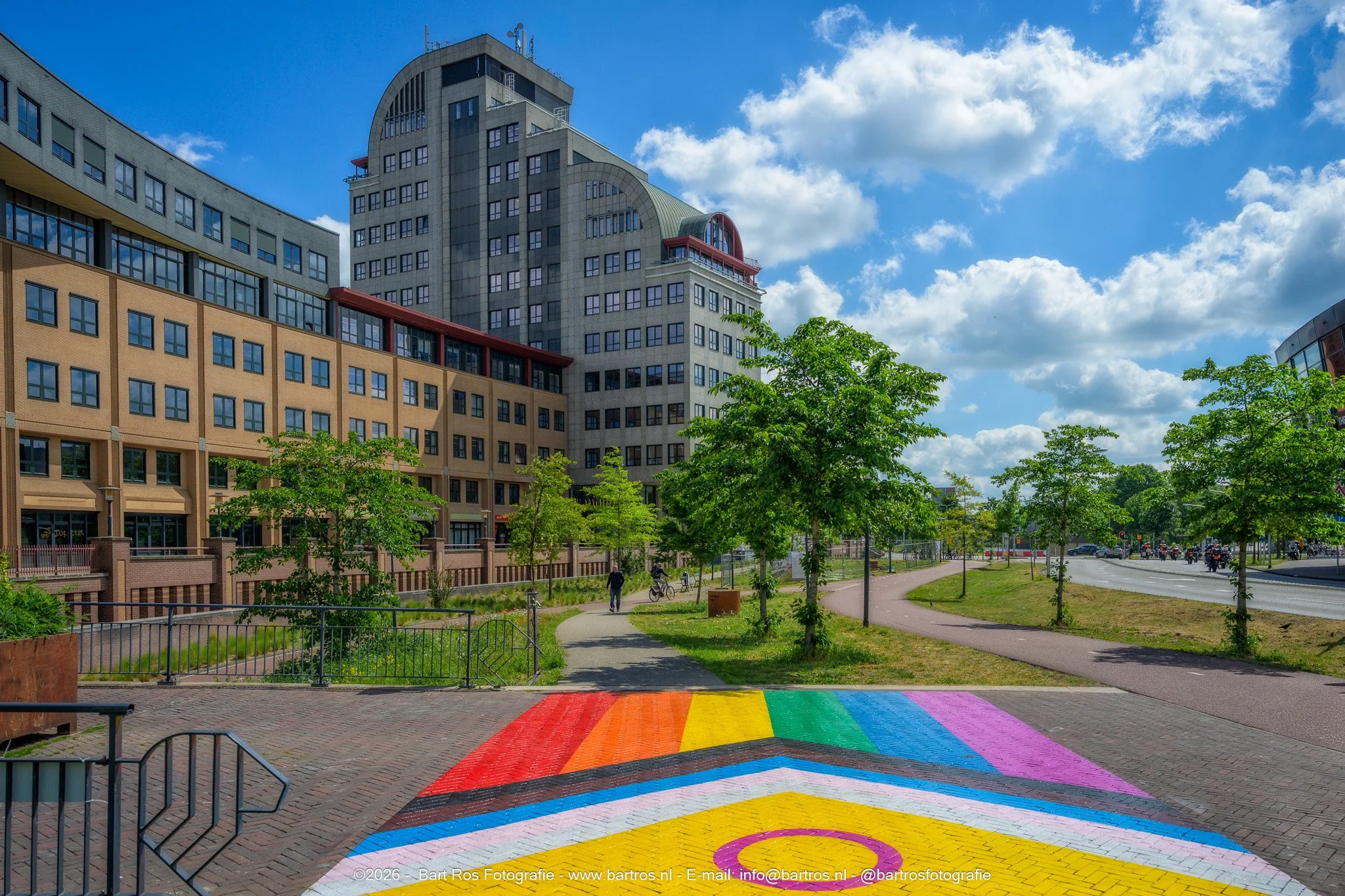 Een stadspark met een kleurrijk zebrapad en een groot modern gebouw op de achtergrond. De lucht is deels bewolkt met blauwe lucht zichtbaar.