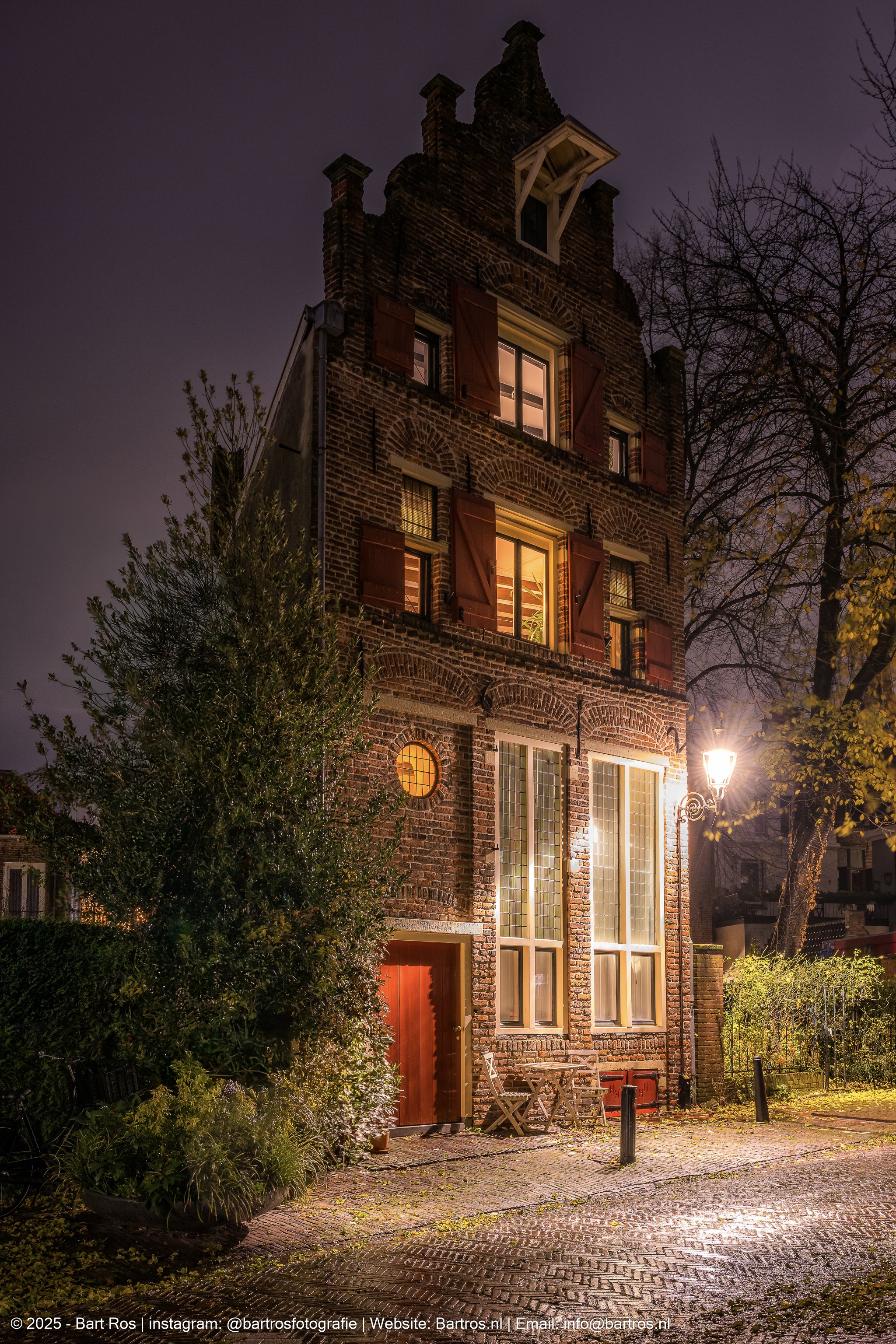 Een oud brick house met open ramen en een open deur, verlicht van binnen, s'avonds met een straatlantaarn en bomen op de achtergrond.