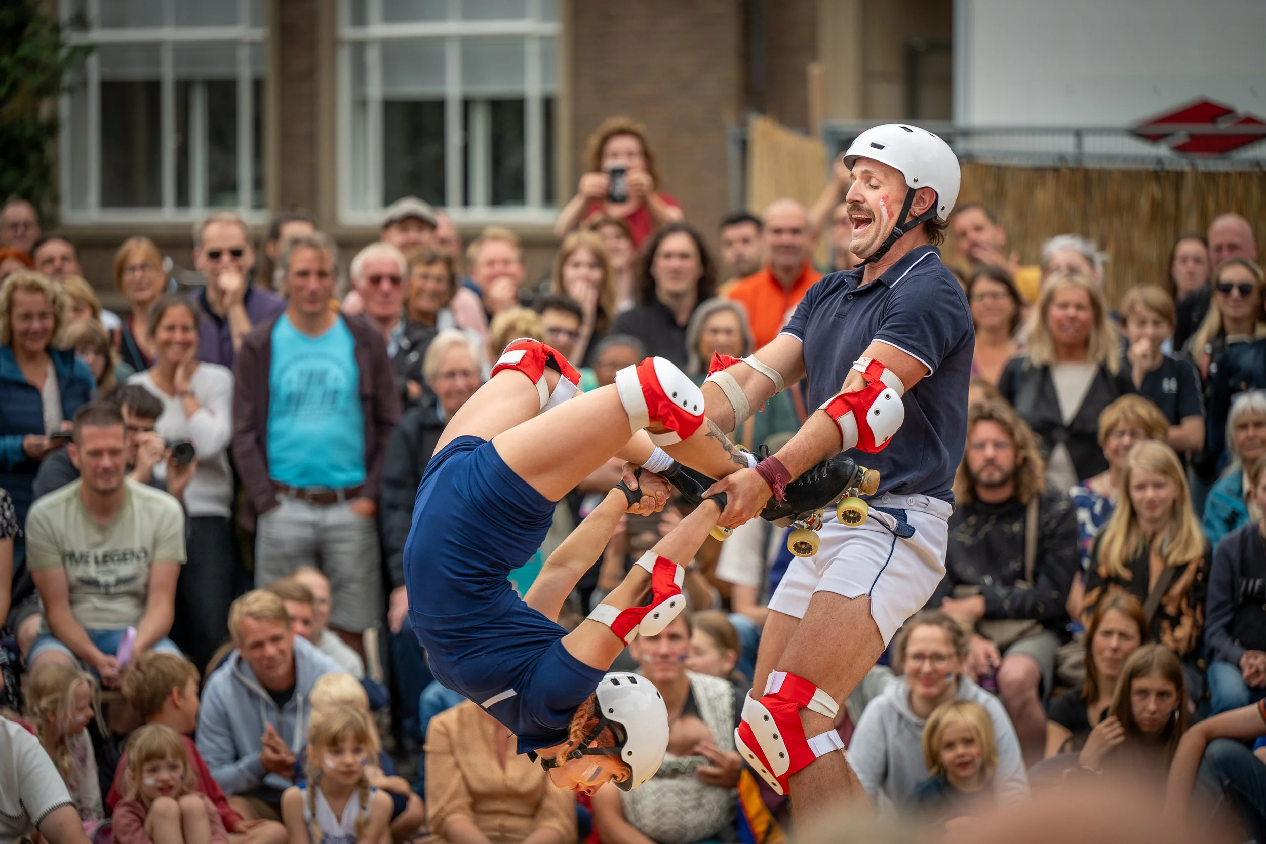 Man en vrouw roller derby deelnemers in uitrusting die samen oefenen voor publiek in een buitenruimte.