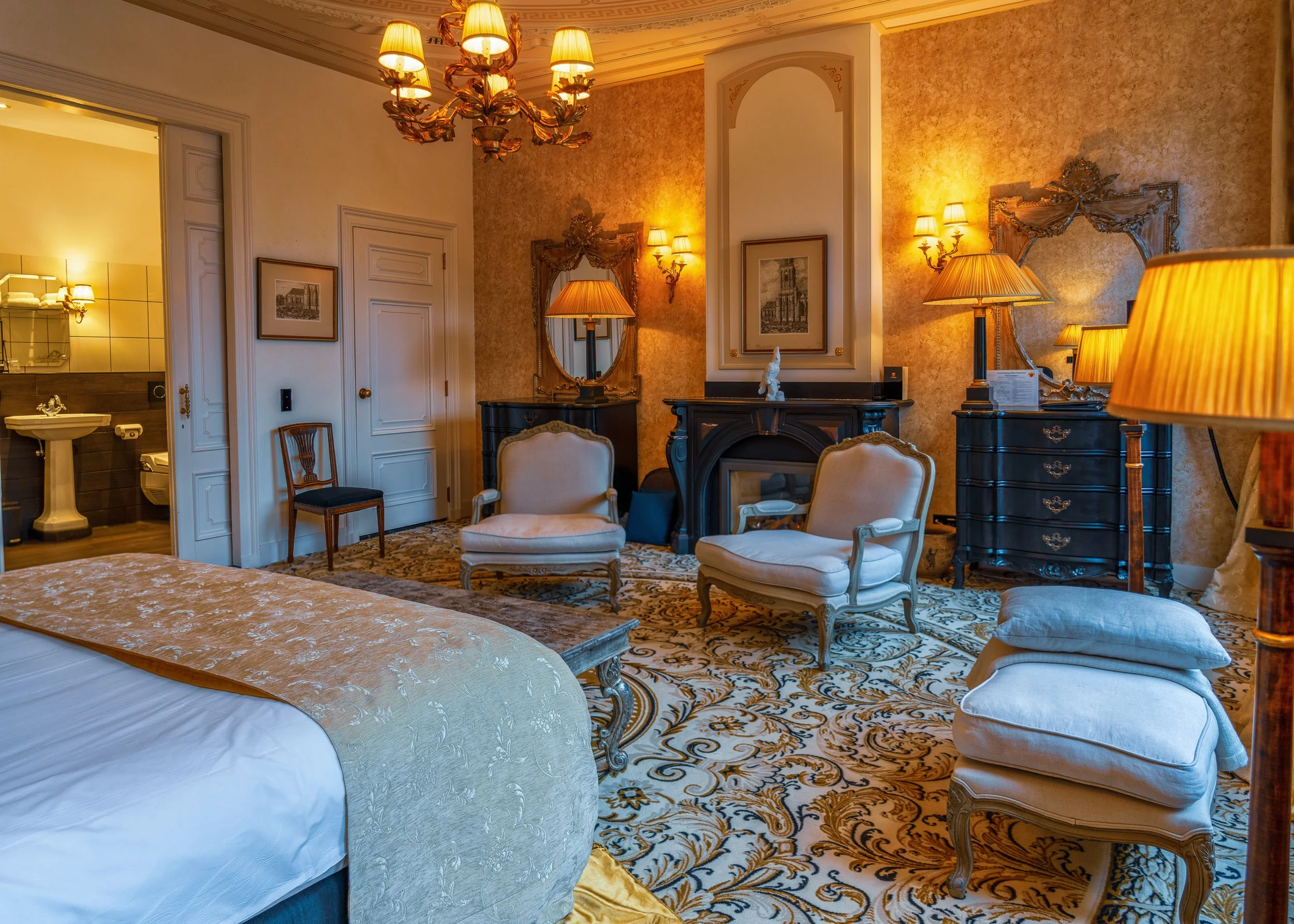 Een luxe hotelkamer met een houten vloer, een antieke kroonluchter, gestoffeerde fauteuils, een zwart dressoir met spiegel, decoratieve schilderijen en sfeervolle verlichting.