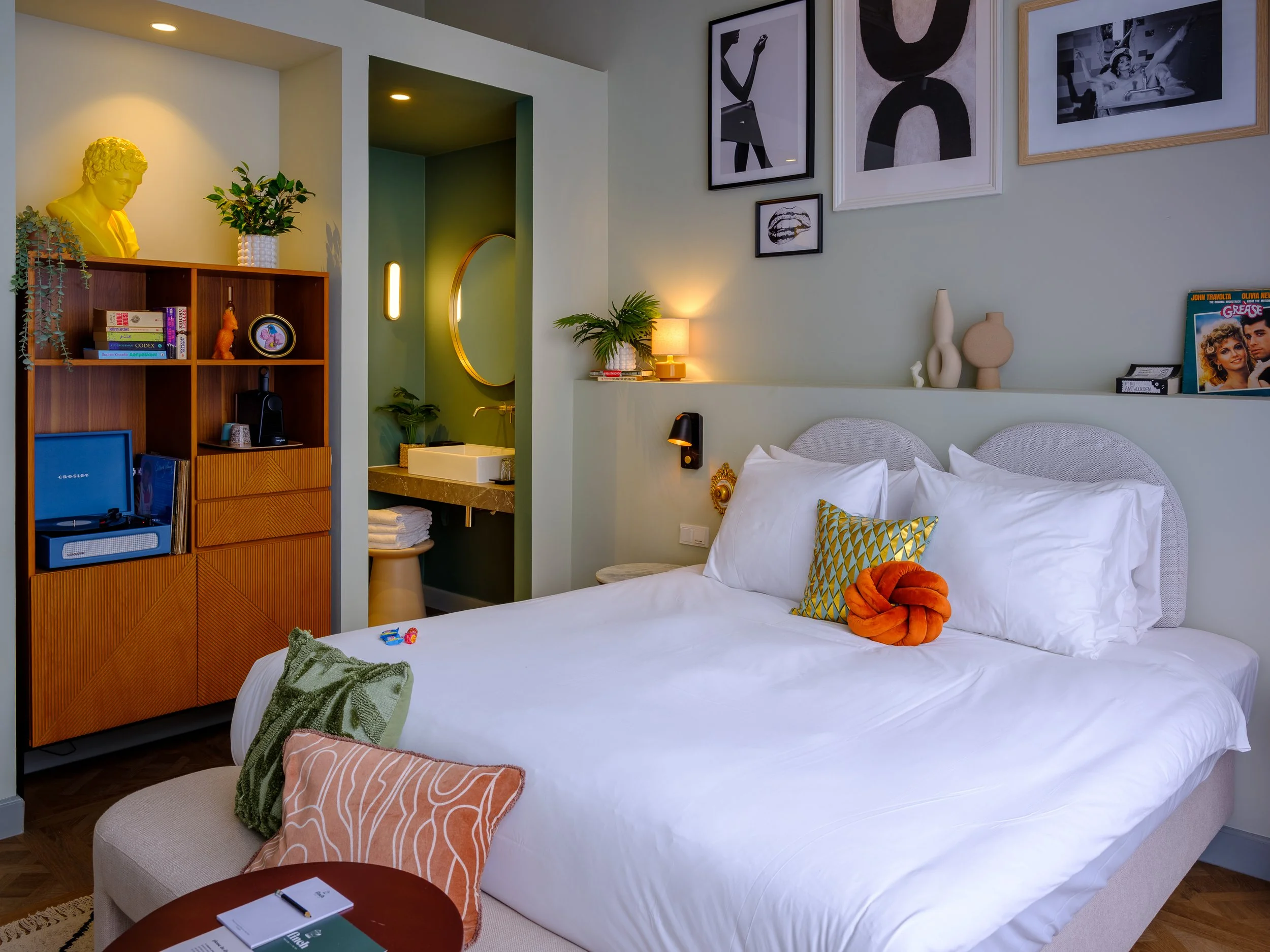 Een moderne slaapkamer met witte bedden en decoratieve kussens, muurkunst en accessoires, een houten boekenkast, en een kleine zithoek met boeken en decoraties.