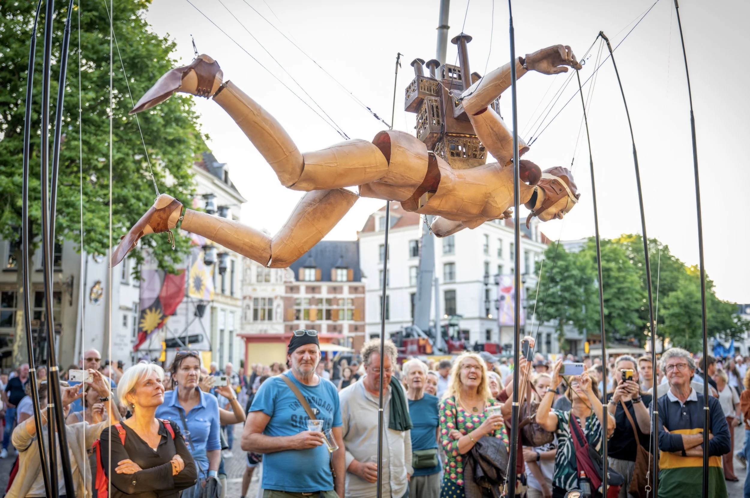 Een grote groep mensen kijkt naar een drijvende roboticaperformance van een vrouwelijke robot in een stad. De robot is opgehangen aan kabels en lijkt te zwemmen of te zweven boven de menigte.