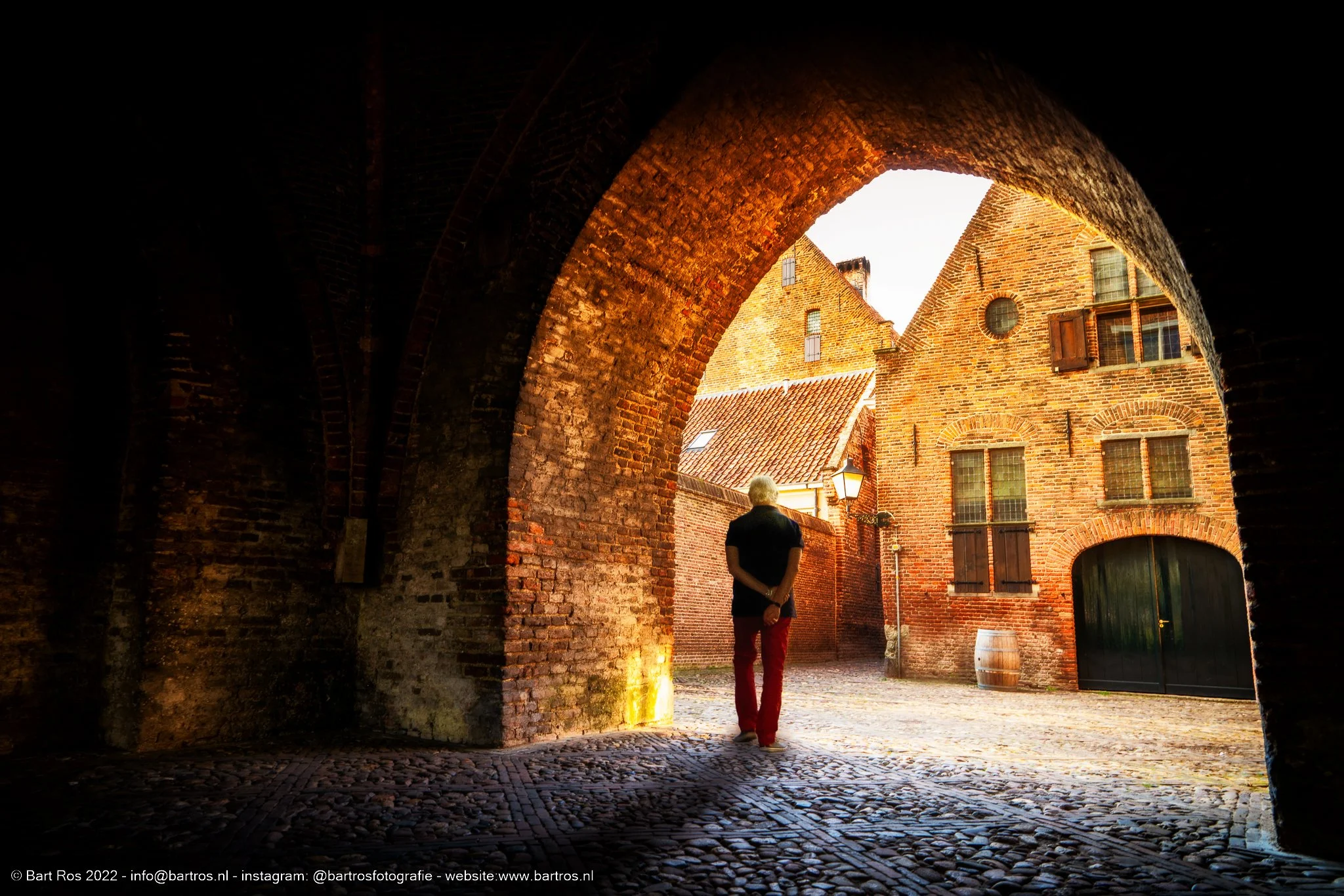 Persoon met witte haren en donkere kleding kijkt uit een ondergrondse poort op een straat met oude bakstenen gebouwen in een historische wijk.