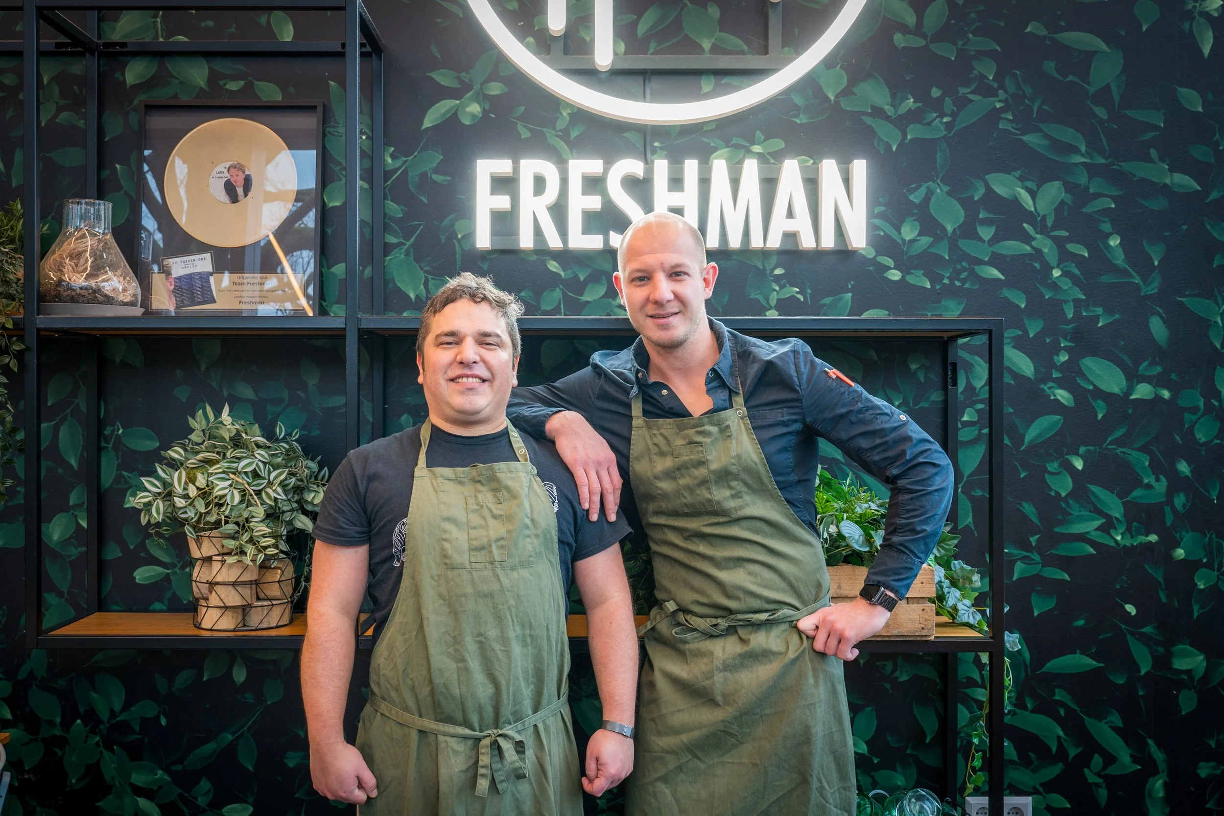 Twee mannen dragen groene aprons en staan in een winkel of restaurant met een donkere, groene, blad-achtige achtergrond en een neonbord dat zegt 'FRESHMAN'.