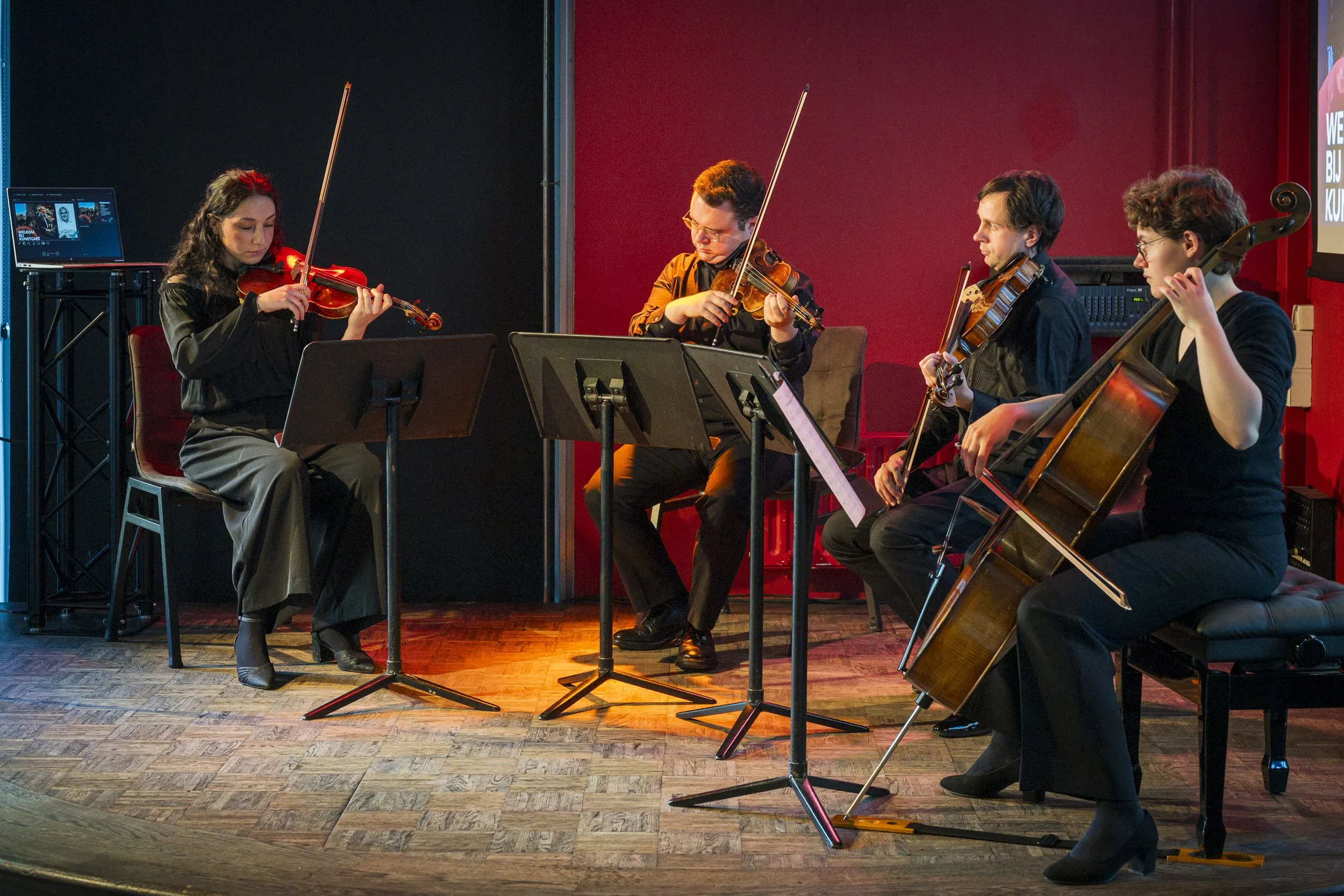 Muzikale kamerorkest bestaande uit twee vioolspelers, een cellospeler en een violist die samen muziek maken, binnen een zaal met een rood en zwart decor.