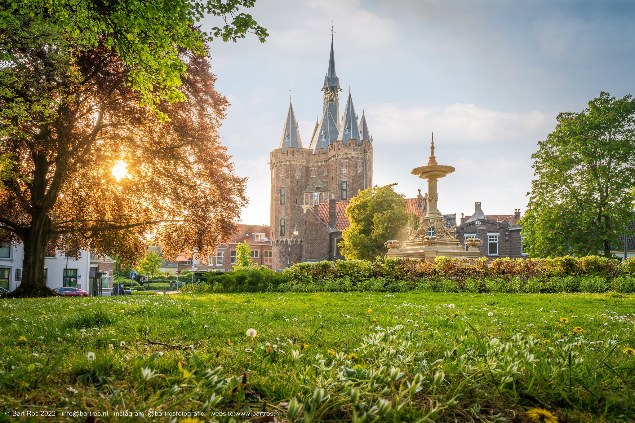 Een tuin met gras, bloemen en bomen, met een fontein en een middeleeuws kasteel met torens op de achtergrond, onder een deels bewolkte blauwe hemel in de namiddag of avond.