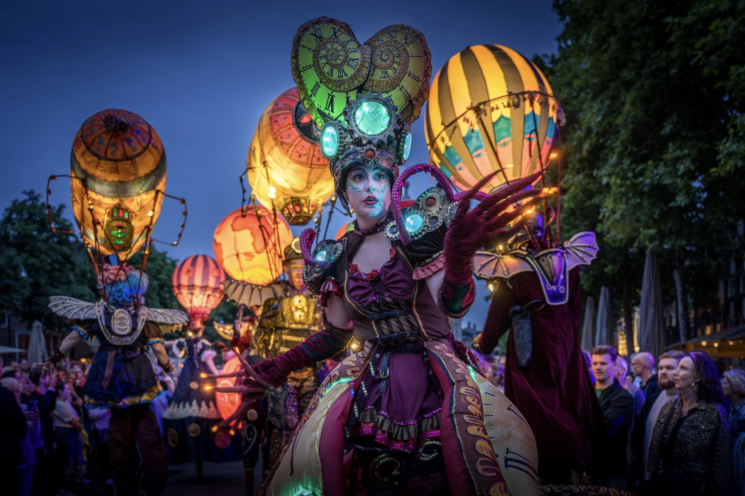Persoon in een kleurrijk fantasy kostuum met verlichte onderdelen, omringd door opblaasbare ballonnen tijdens een parade in de avond.