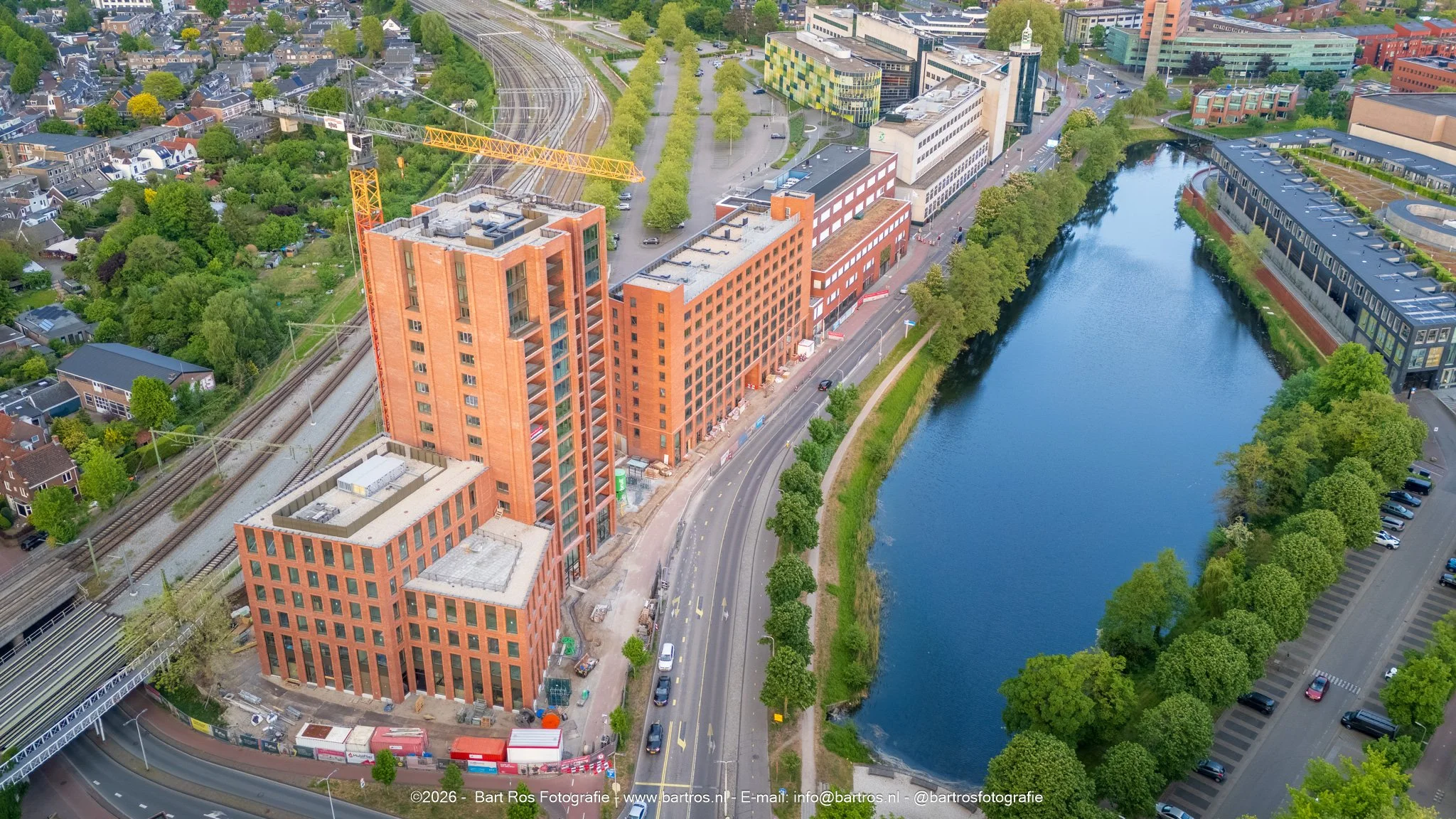 Een hoog gebouw onder constructie langs een rivier, met een bouwkraan en een spoorlijn, omliggende woningen en moderne architectuur, groene bomen en een waterkant.