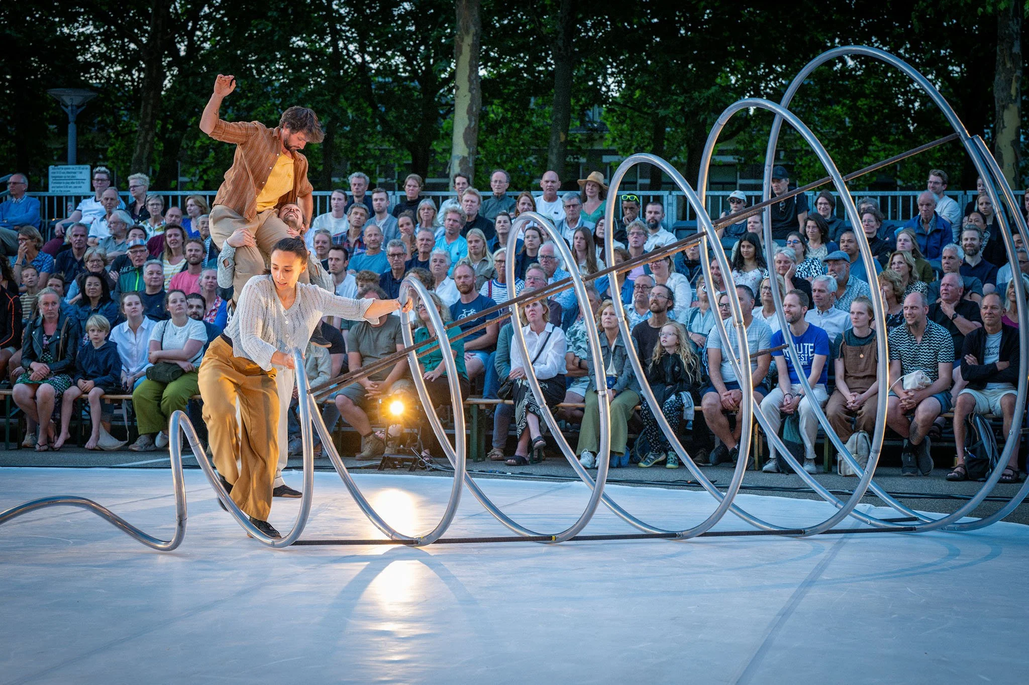 Twee performers op een podium met een publiek zittend in de achtergrond, bij avondlicht, onderdeel van een show of theater optreden.