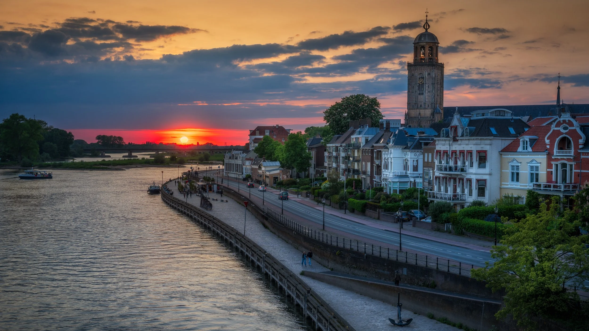 De rivier de IJssel in Deventer met een promenade en huizen langs de oever, onder een kleurrijke zonsondergang met een bewolkte hemel.