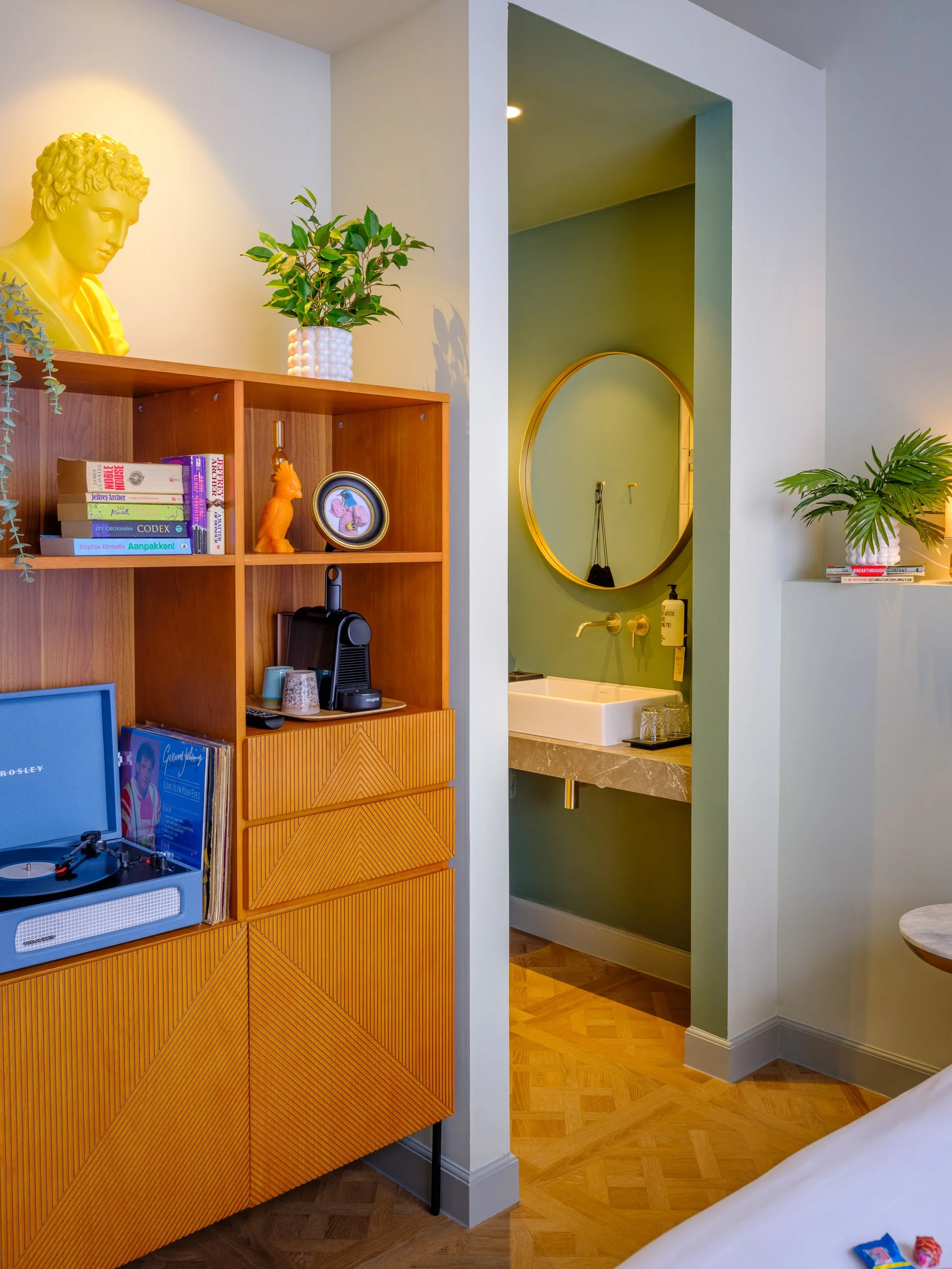 Interieur van een kamer met een houten boekenkast, een groene plantenbak en een spiegel in een nis. In de turquoise nis bevindt zich een wasbak en een zeepdispenser. Op de boekenkast staan boeken, een vaas, en een stereo met platen.