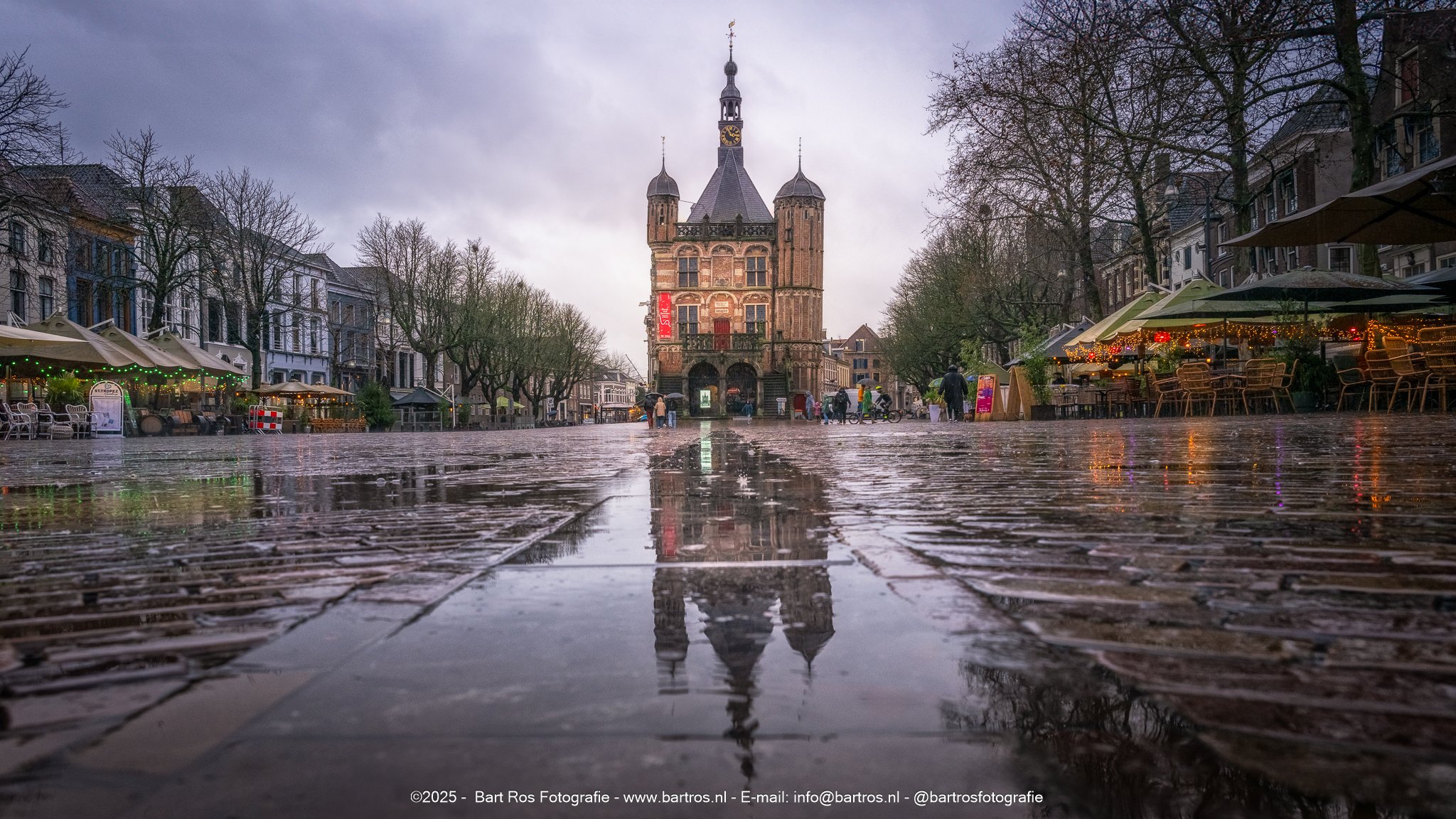 Een oude kerk met een toren en een wijzerplaat, omringd door rijen bomen en gezellig uitziende cafés met verlichting. De straat ligt nat, mogelijk na regen.