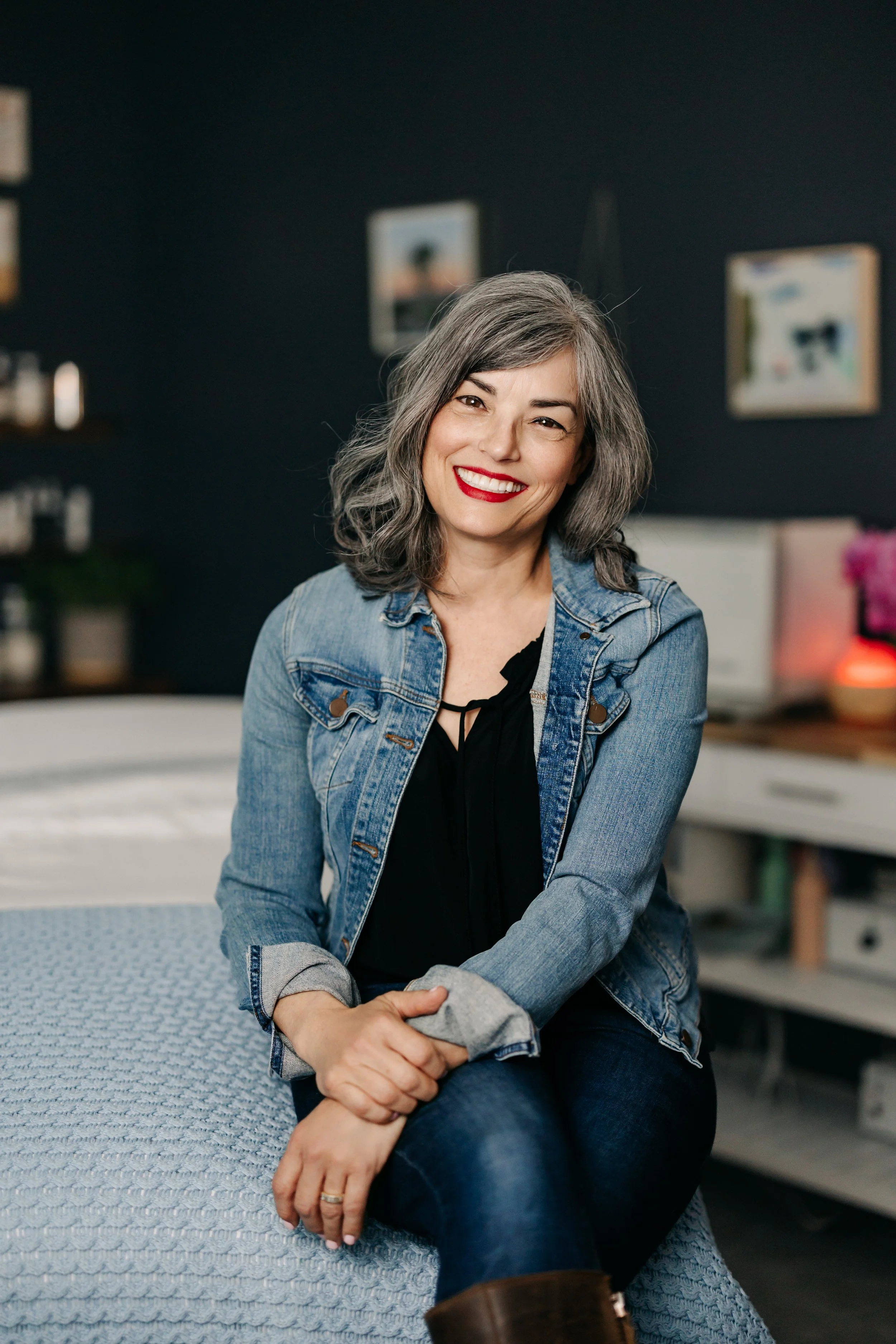 A smiling woman with gray hair, wearing a denim jacket and dark jeans, seated on a textured blue chair in a room with dark walls and framed pictures in the background.