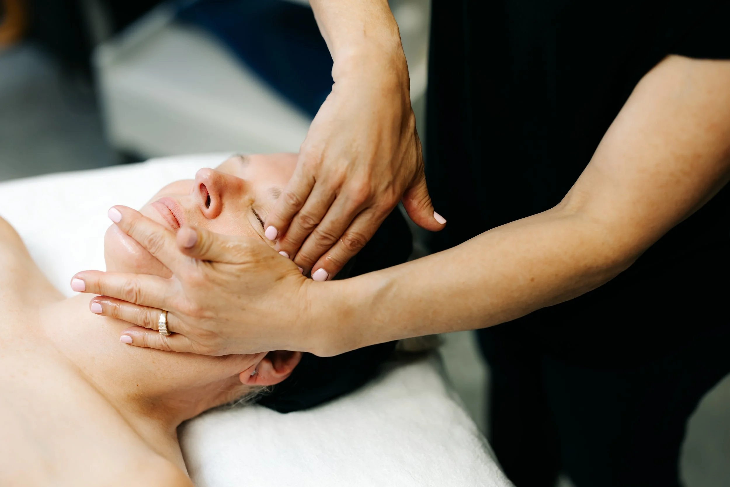 A woman receives a massage in a spa, with a therapist using her thumbs on the woman's face while the woman lies face-up with eyes closed.