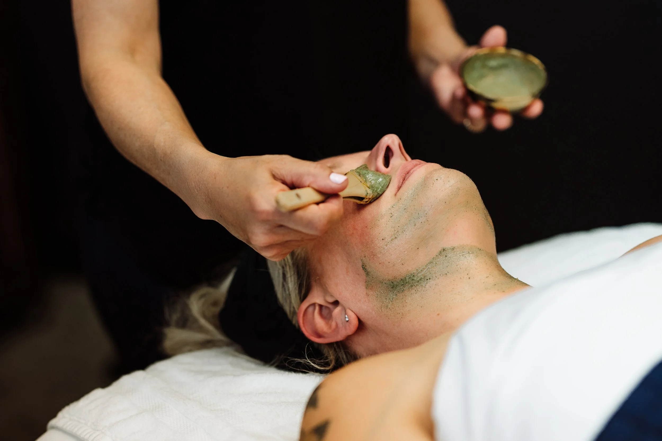 A woman receiving a facial treatment with a green mask, lying down with her eyes closed, while an esthetician applies a green substance to her face using a brush.