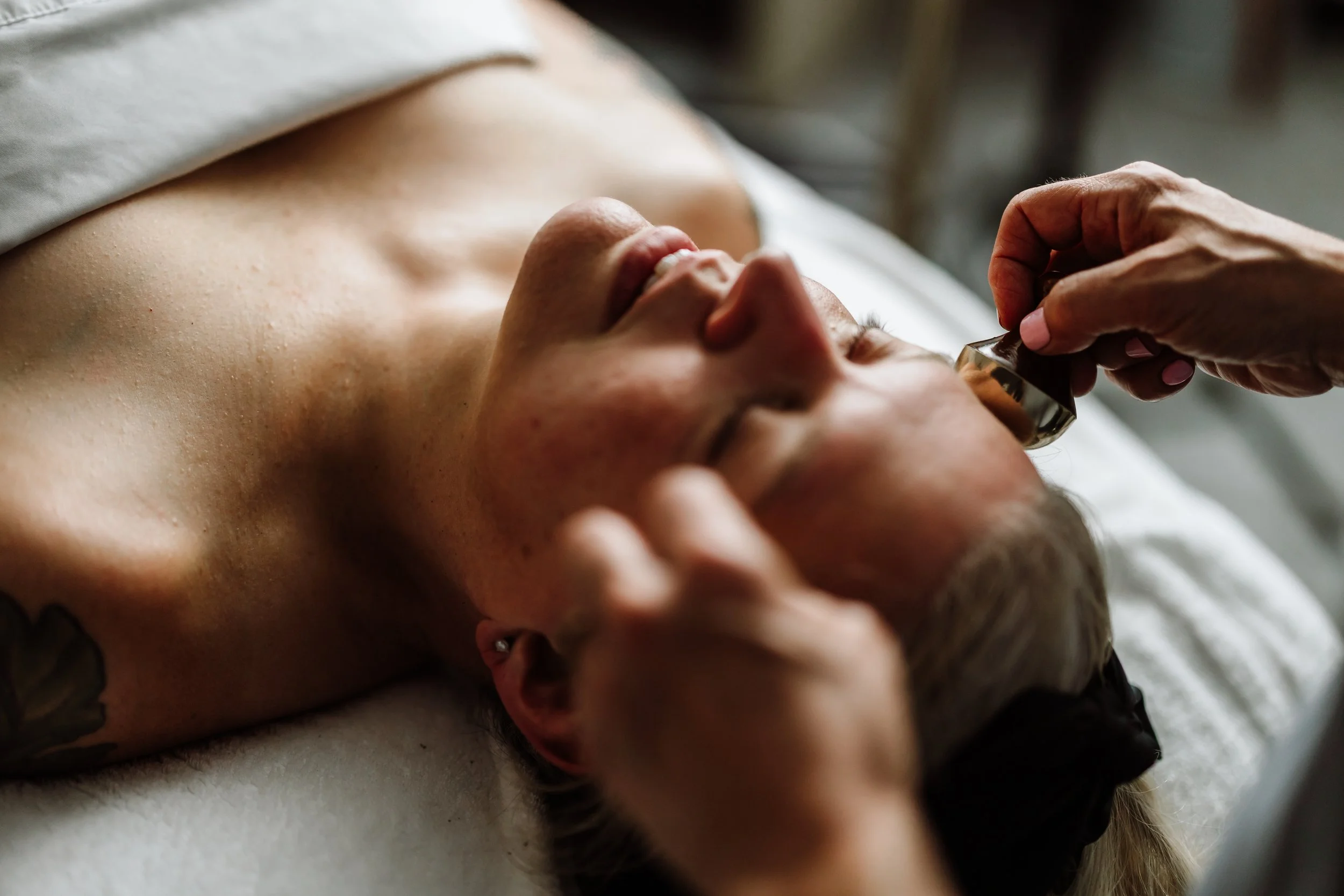 A person receiving a facial, lying down with their eyes closed and are very relaxed, while the esthetician is using their hands for the treatment.