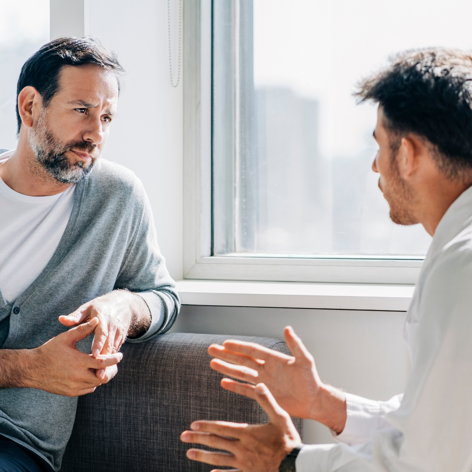 Man with gray beard and gray sweater listening and talking to a younger man in white shirt in a bright room near a window.