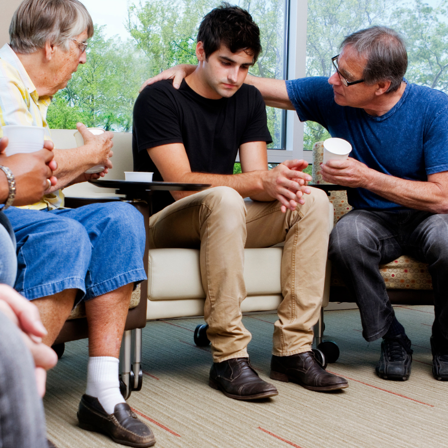 A young man sitting on a couch with two older men, one on each side, comforting him during a conversation. All three are holding cups, and the background shows a large window with green trees outside.