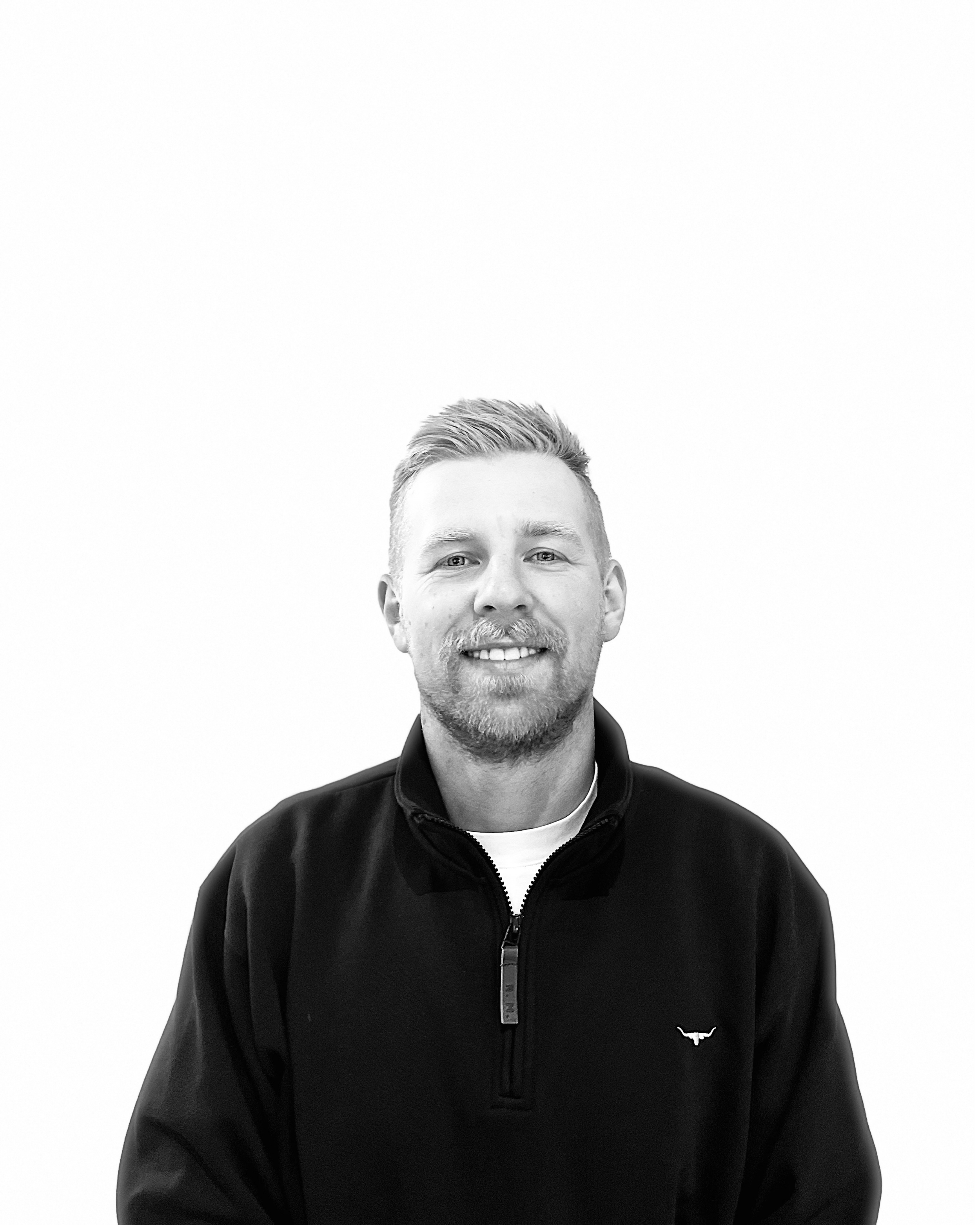 A smiling man with short hair and a beard, wearing a black jacket with a zipper and a small logo on the chest, against a plain white background.