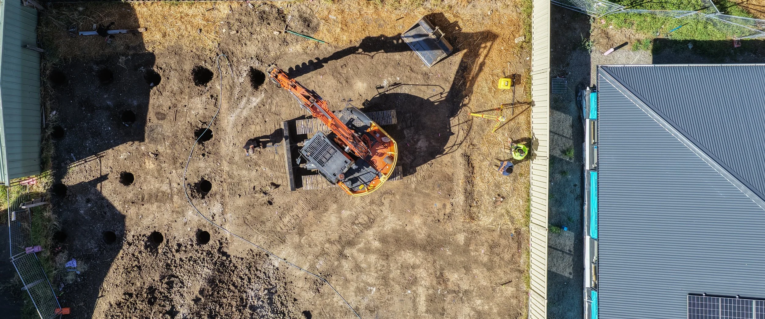 An aerial view of a construction site featuring an orange excavator, two construction workers, and various tools. The site is adjacent to a metal building with a gray roof and a white fence.