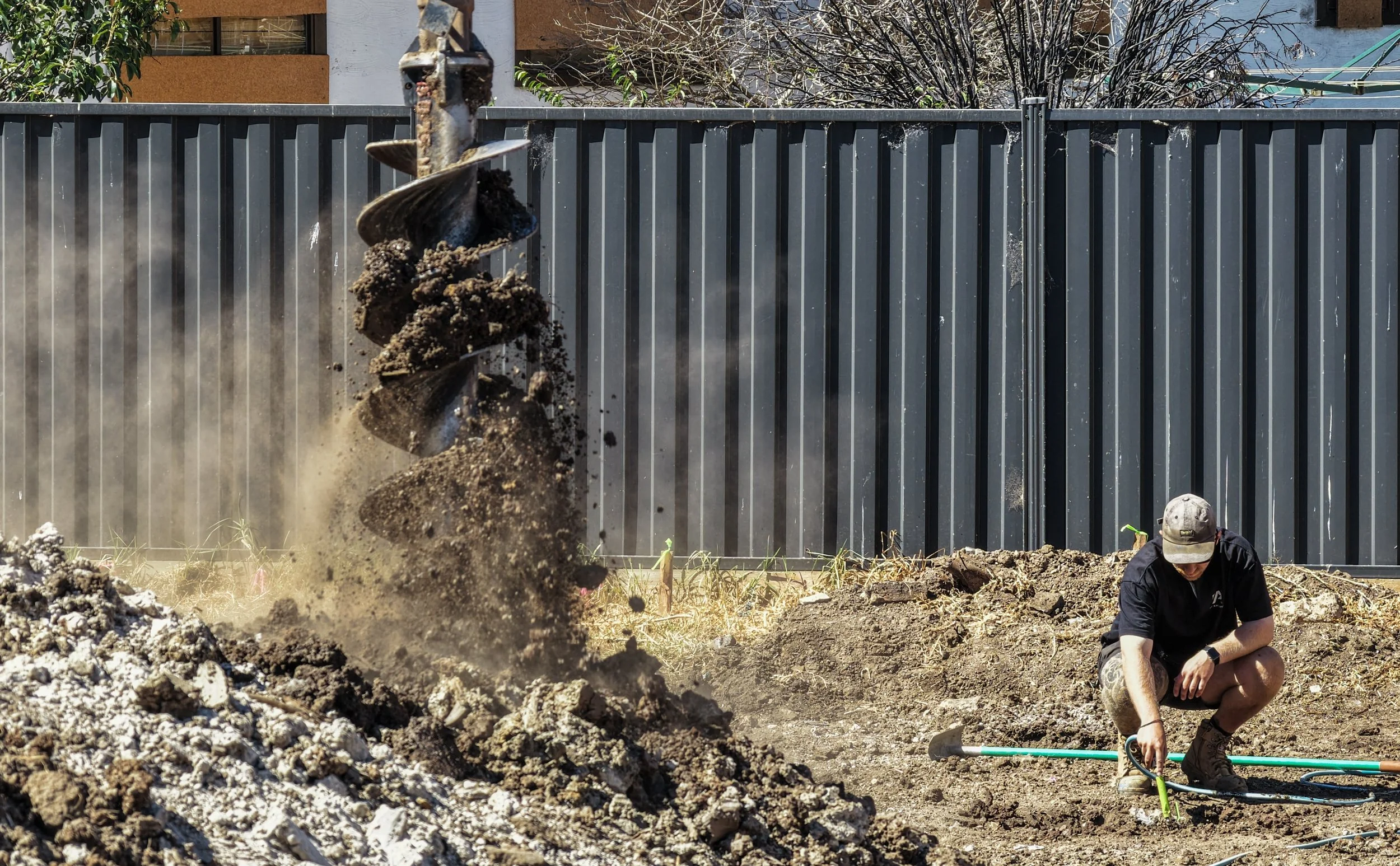 A person wearing a baseball cap working on a construction site with a large metal fence in the background and a construction drill in their hands. A pile of dirt is in the foreground, with dust and dirt flying in the air.