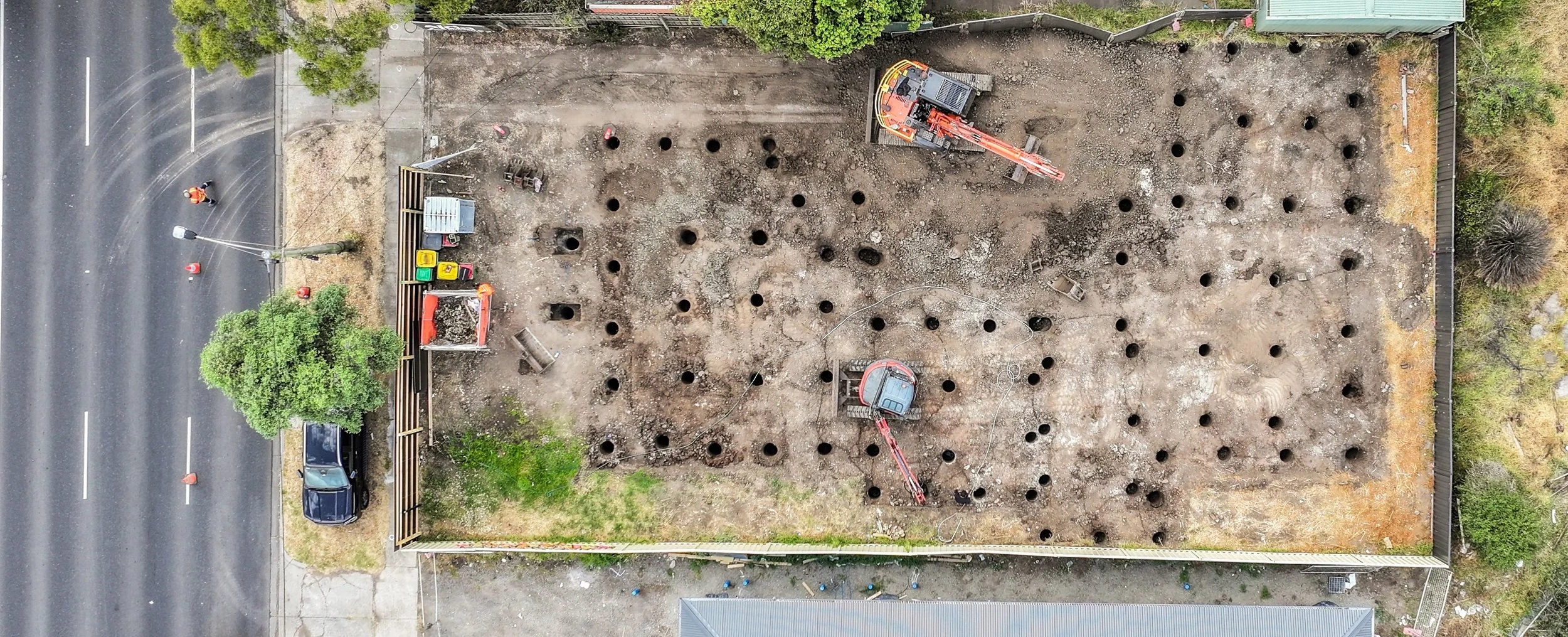 Construction site with holes in the ground, two excavators working, and equipment along the perimeter.