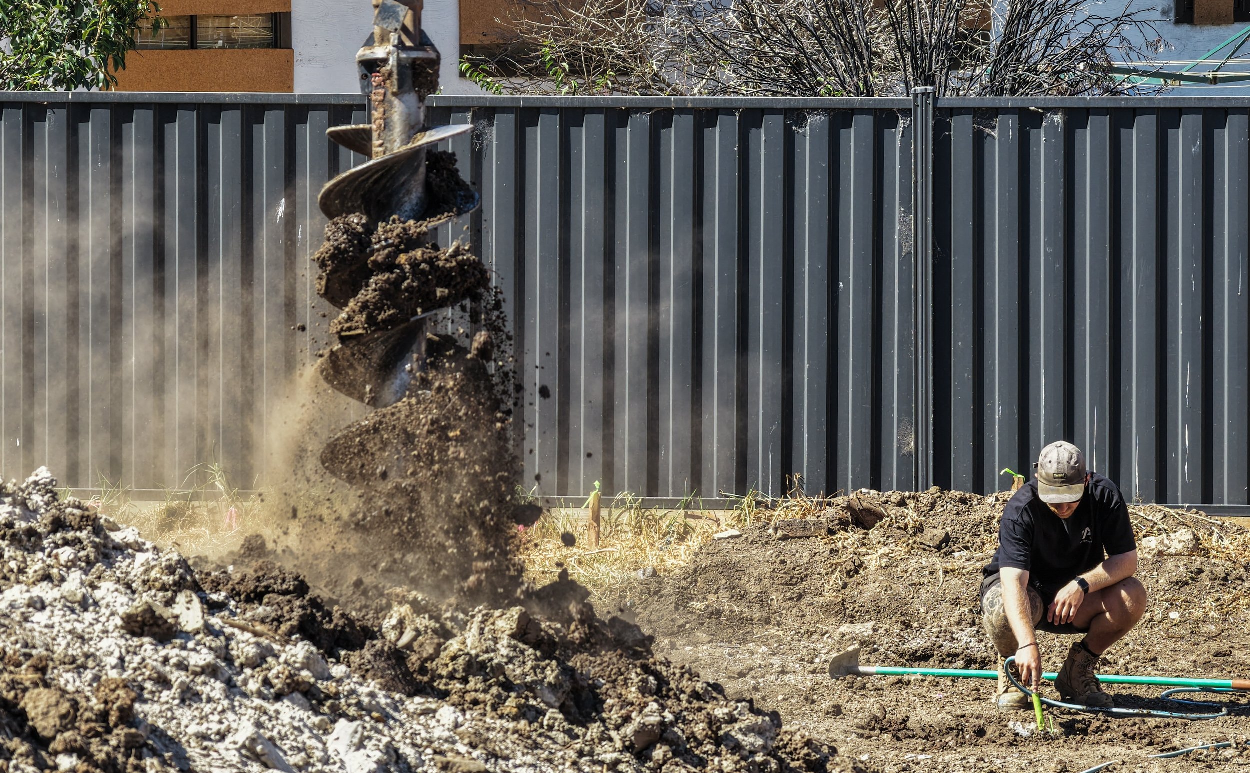 A construction worker wearing a cap is kneeling on the ground, working with tools amidst dirt and soil at a construction site. In the background, a grey metal fence and some trees are visible.