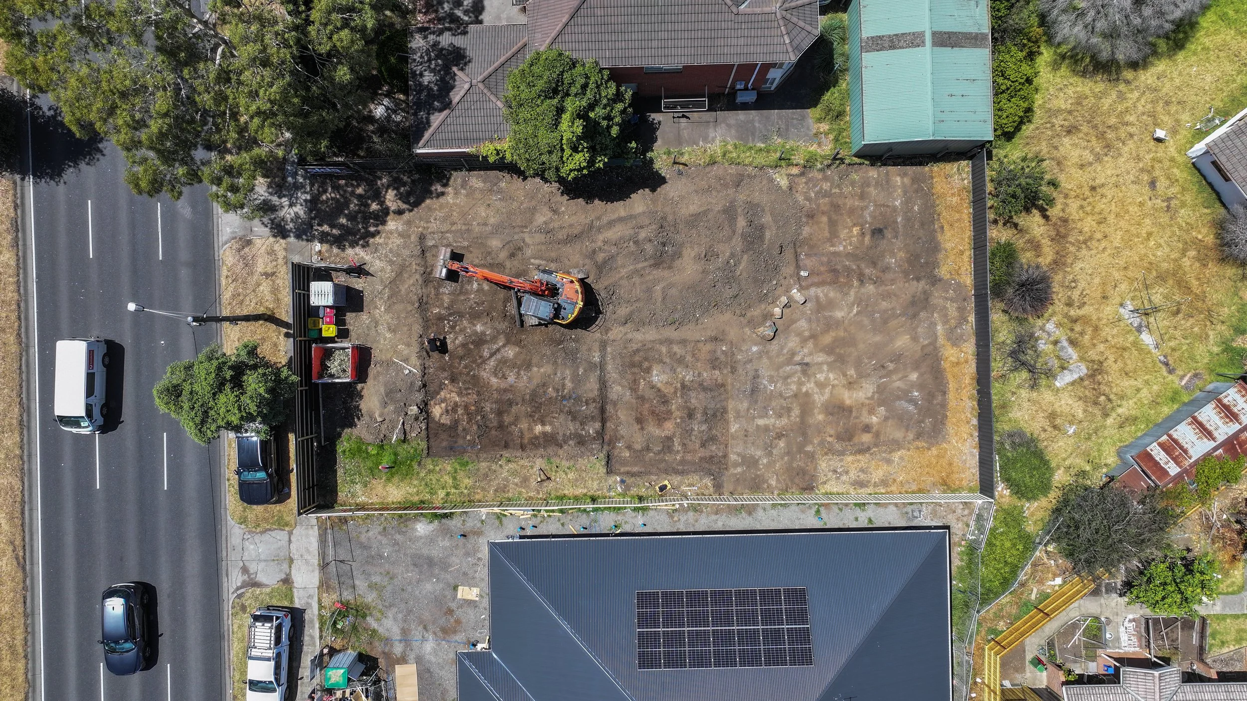An aerial view of a construction site with a backhoe, surrounded by partially excavated dirt, adjacent to a street with parked cars and neighboring buildings with trees and gardens.