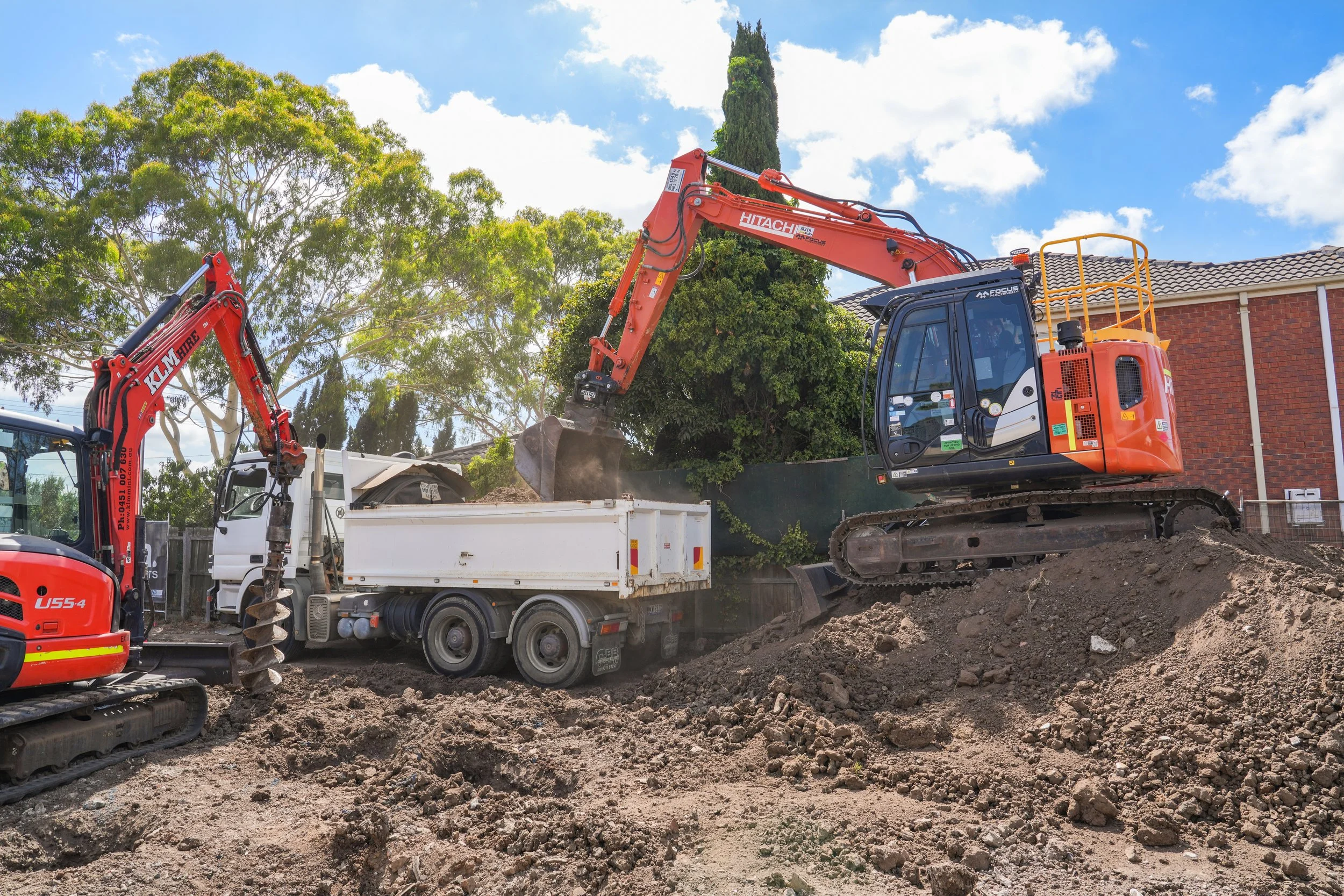 Construction site with an orange excavator scooping dirt into a white truck, other construction equipment visible, residential house in the background, trees, and a partly cloudy sky.