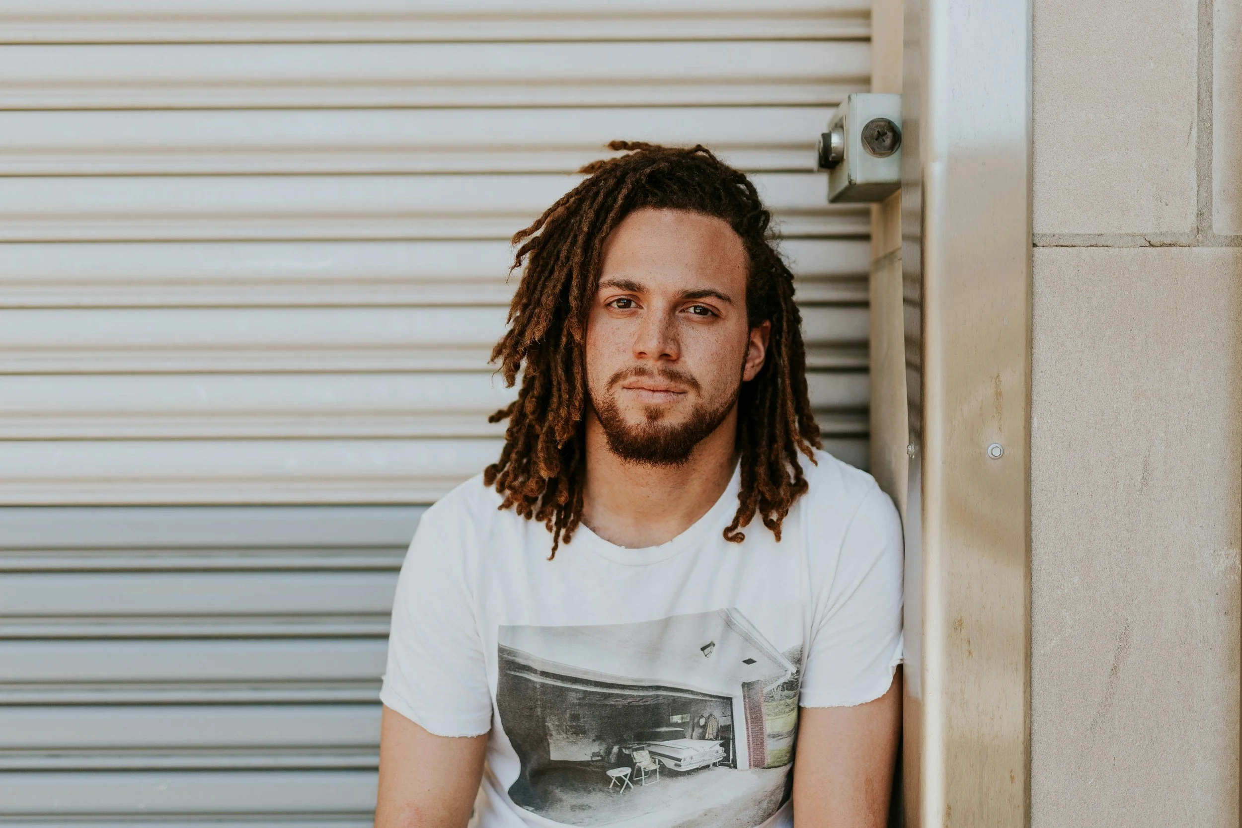 A man with dreadlocks and a beard sitting in front of a metal shutter and a beige wall, wearing a white t-shirt with a graphic image on it.baog