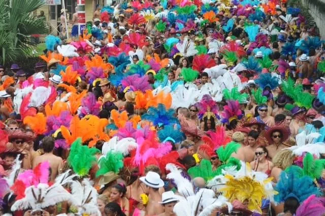 Crowd of people participating in a colorful parade or festival, many wearing vibrant feathered costumes and hats.baog