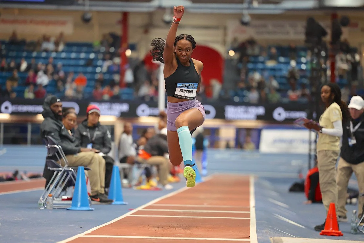 A female athlete jumping in a long jump event at an indoor track and field competition.
