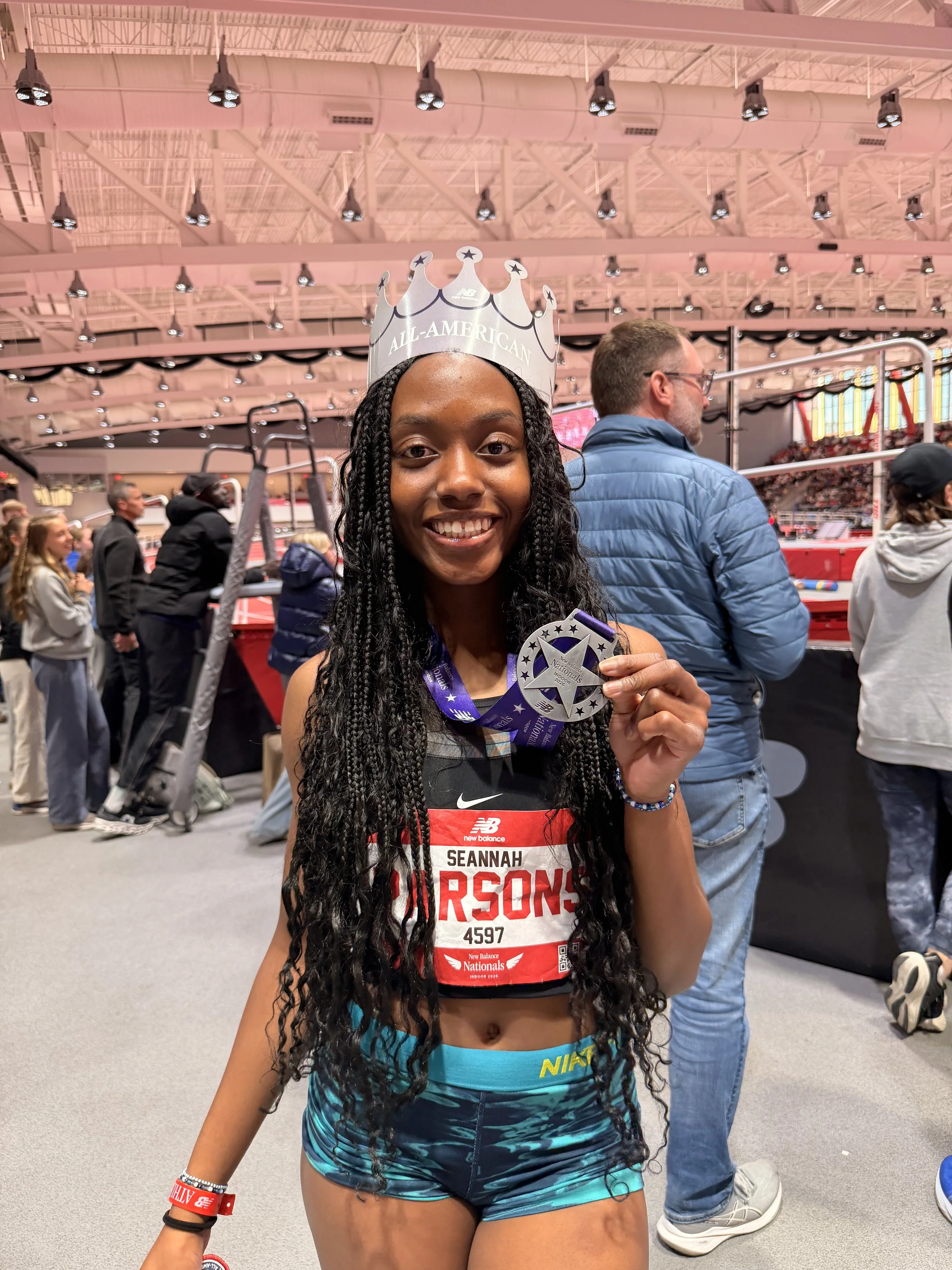 A young woman with dark curly hair wearing sportswear, a crown that says 'All-American', and holding a medal, smiling at a race event.