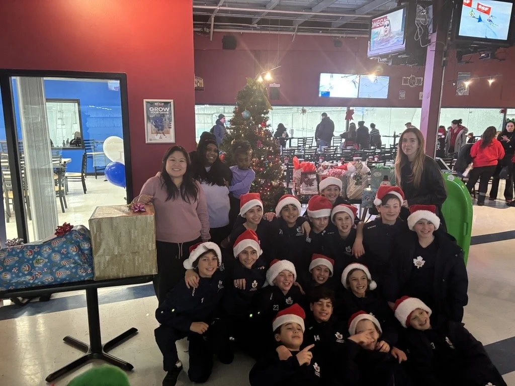 Group of children and adults celebrating Christmas with a decorated tree, wearing Santa hats, in an indoor setting with holiday decorations.