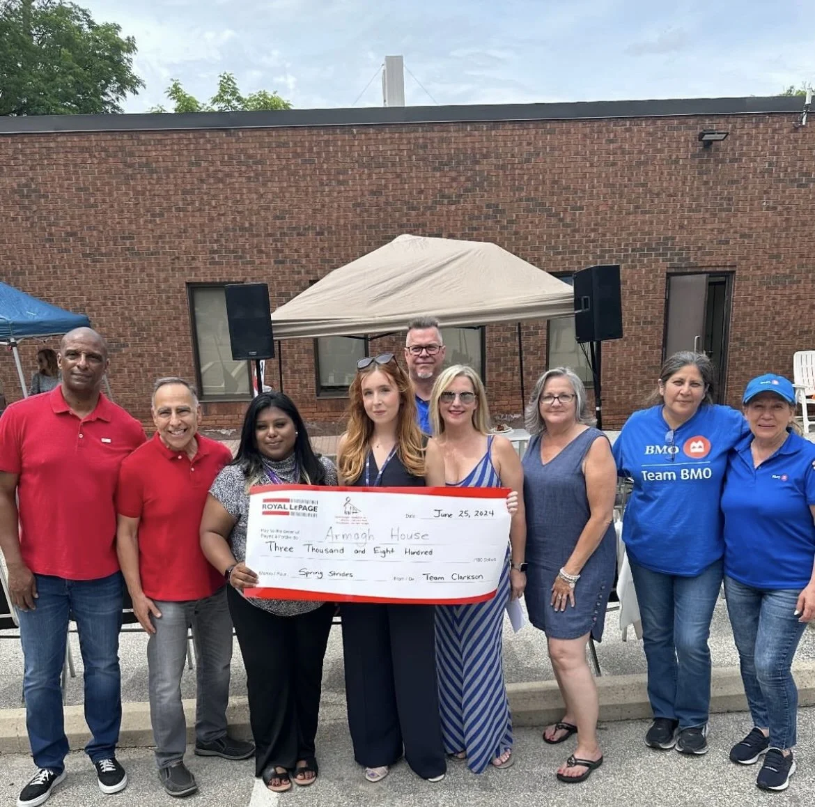 Group of people holding a large ceremonial check outdoors during daytime, standing in front of a brick building with tents and speakers set up.