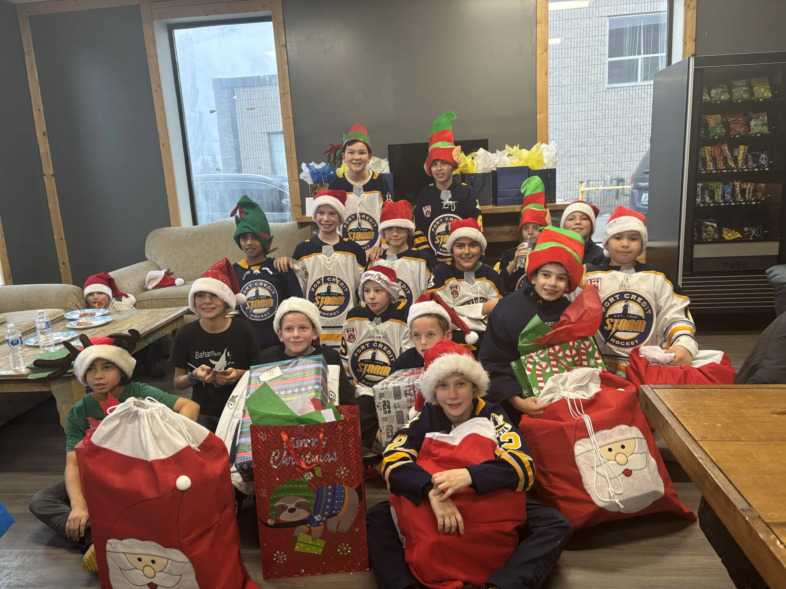A group of children wearing Santa hats and elf hats inside a room, smiling and gathered around Christmas gift bags, with a vending machine in the background.