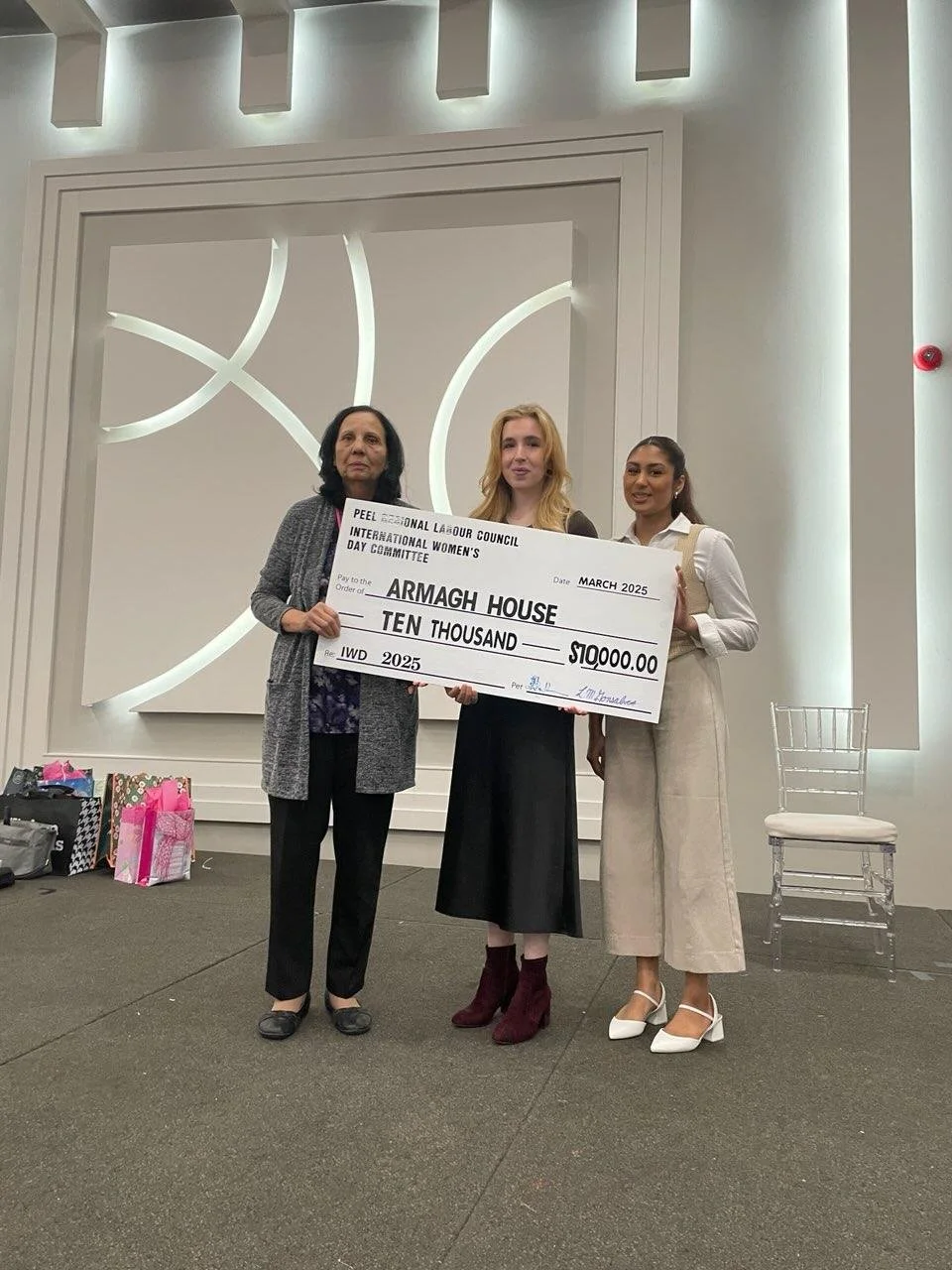 Three women standing in a room holding an oversized check for ten thousand dollars. The check is addressed to ARMAGH HOUSE and dated March 2025. The women are dressed in formal attire, and there are gift bags and a chair in the background.