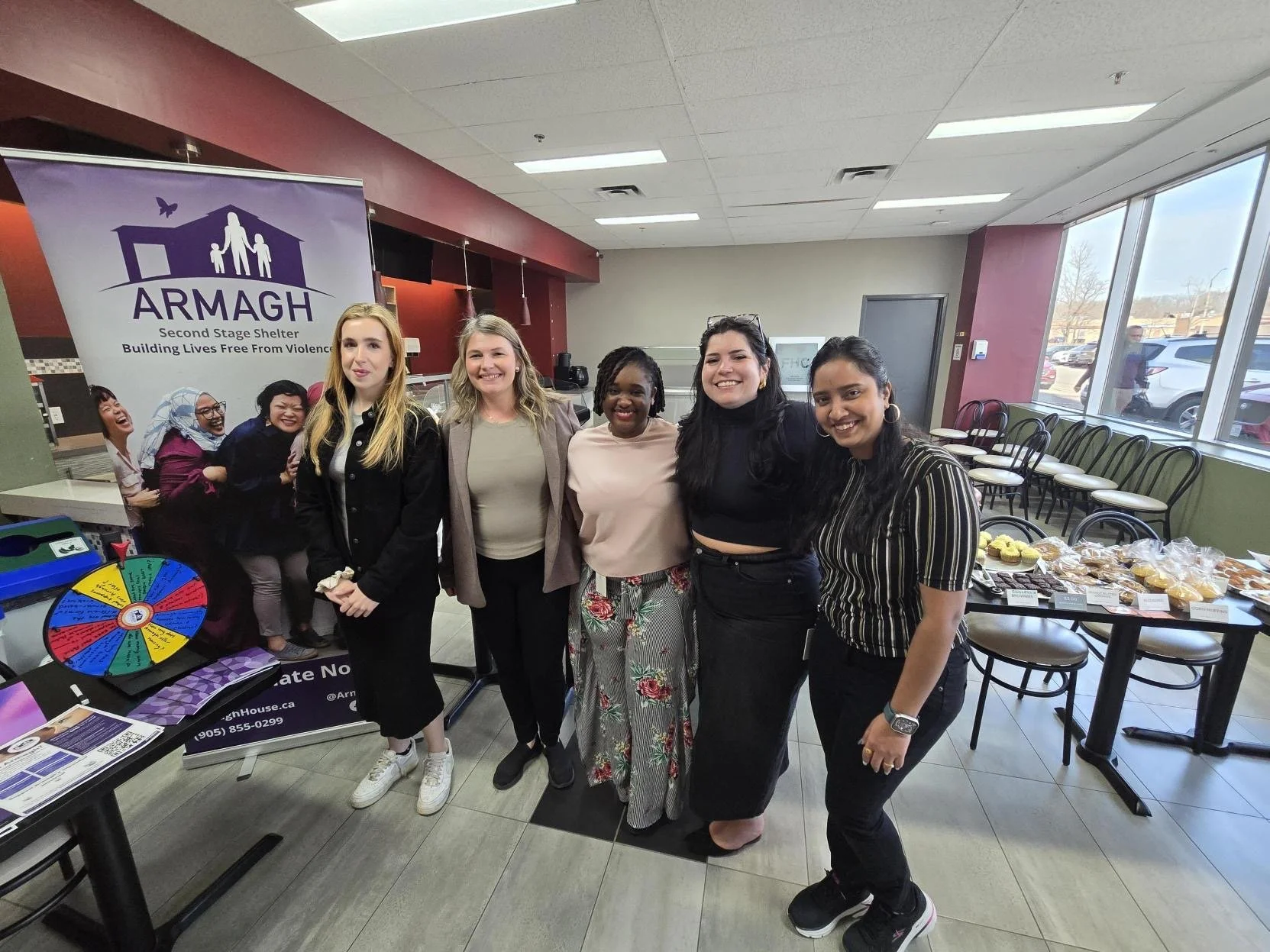 A group of six women is posing inside a building near a counter with a spinning prize wheel, a table with baked goods, and a banner for ARMAGH, an organization focused on building lives free from violence. The women are smiling and standing close together.