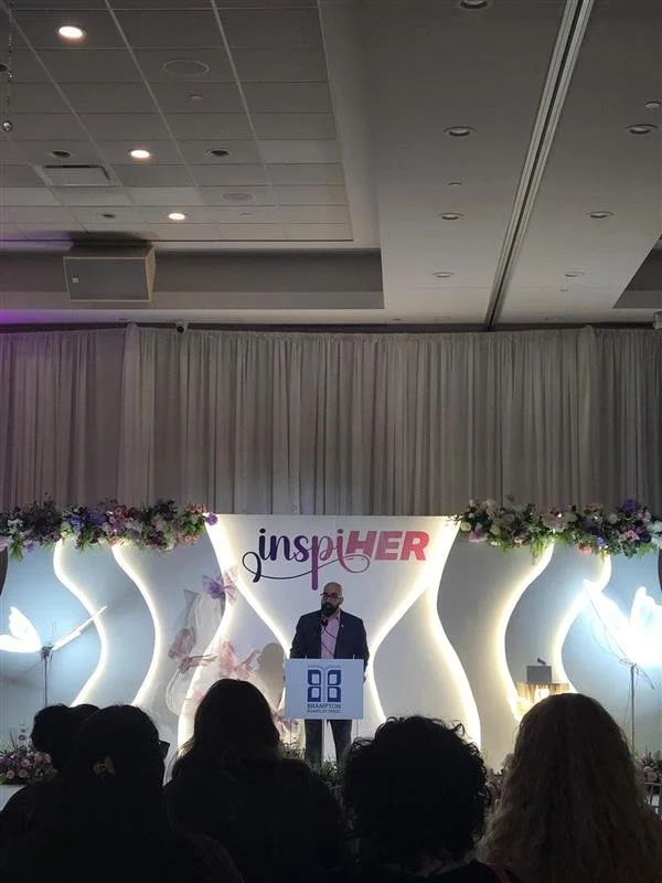 Man in a suit speaking at a podium during an event called 'inspiHER.' The backdrop features a floral decoration and a large sign with the event's name, with women and butterfly images and floral arrangements on the stage.