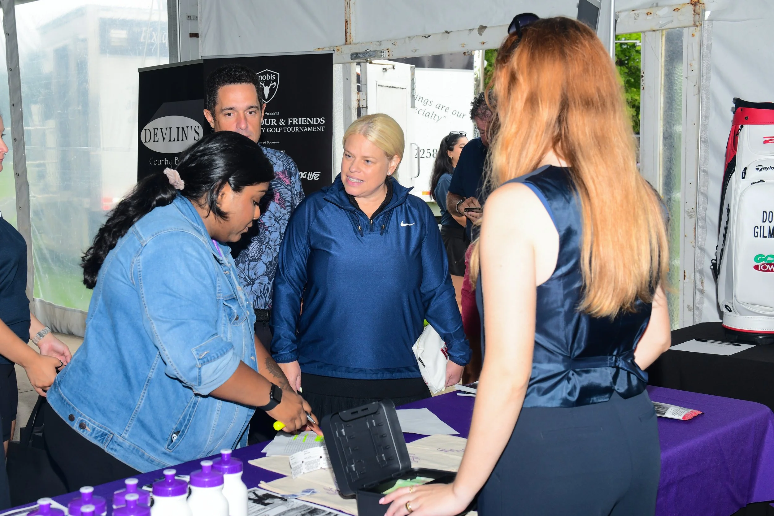 Several women gathered around a table with purple bottles, engaging in an activity at an event inside a tent with banners in the background.